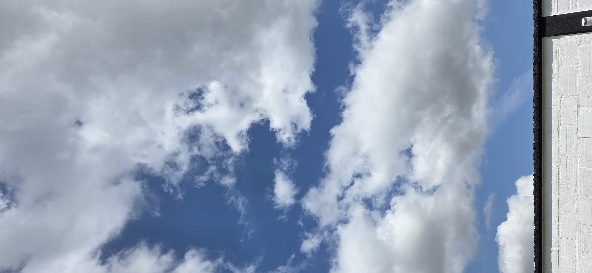 The image depicts a cloudy sky with some blue patches visible. The clouds are white and puffy, indicating fair weather. To the right of the image, a white brick wall with black trim is visible, suggesting an outdoor setting. The perspective is from below, looking up at the sky. No people or specific objects are discernible beyond the clouds and the wall. The lighting suggests it is daytime, and the weather appears to be partly cloudy. There is no visible text in the image. The overall scene is tranquil and natural.
