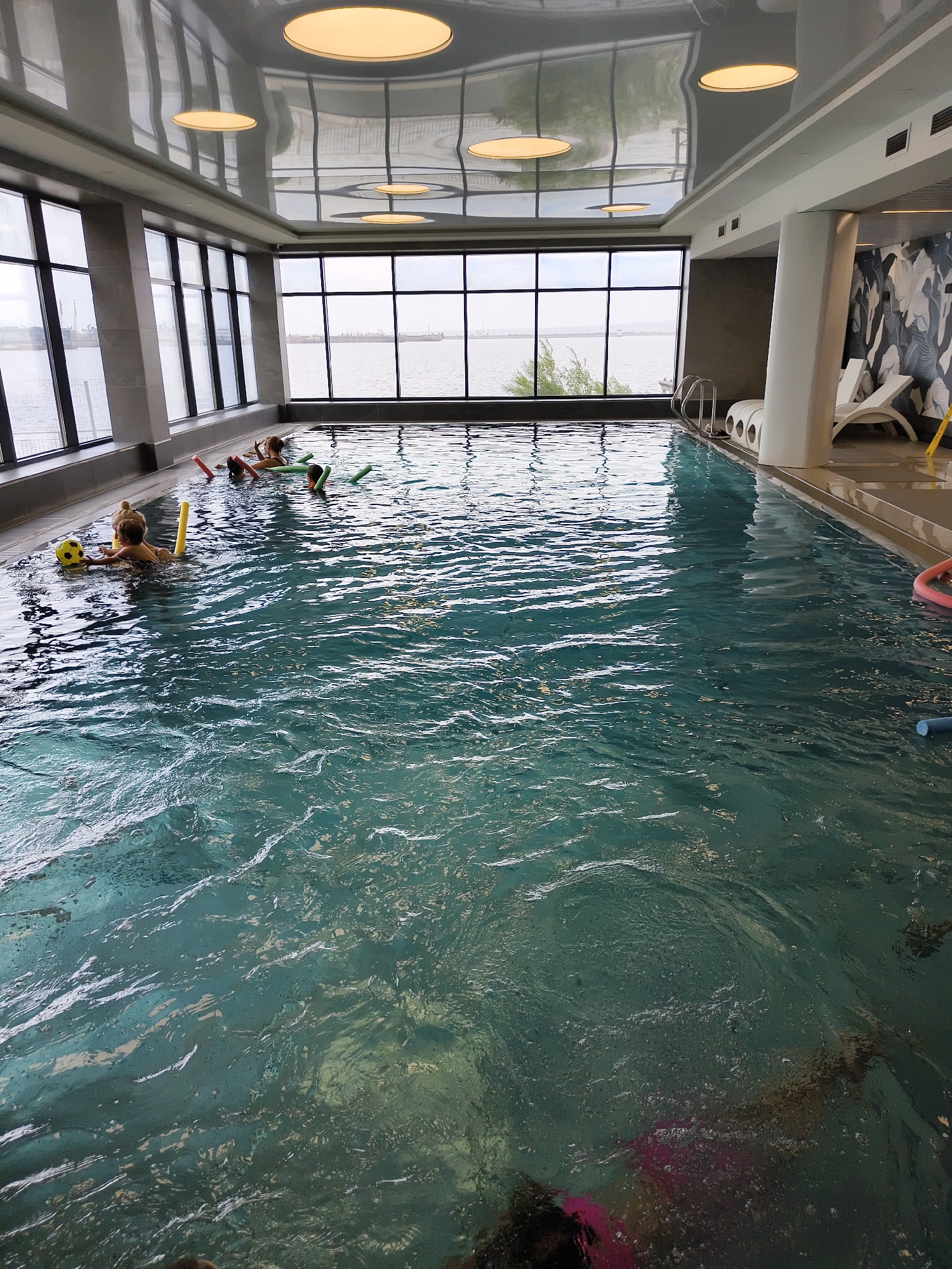 In this indoor swimming pool, several children are engaged in water activities. The pool is surrounded by large windows offering a view of a body of water and the sky, suggesting it could be daytime. The ceiling features modern, circular light fixtures.
The main subjects are children in the water, some holding onto pool noodles and a yellow ball. One child, a young girl, is seen holding the yellow ball and has a pool noodle. Others are interacting with noodles. One child is partially submerged, wearing a pink swimsuit. The water is clear and a deep blue-green, with ripples on the surface.
The setting appears to be a resort or a spa, given the modern design, lounge chairs visible on the side, and the large windows. The light reflecting off the ceiling and water creates a bright, airy atmosphere. The weather outside appears overcast, as indicated by the diffused light coming through the windows. There is no visible text in the image.