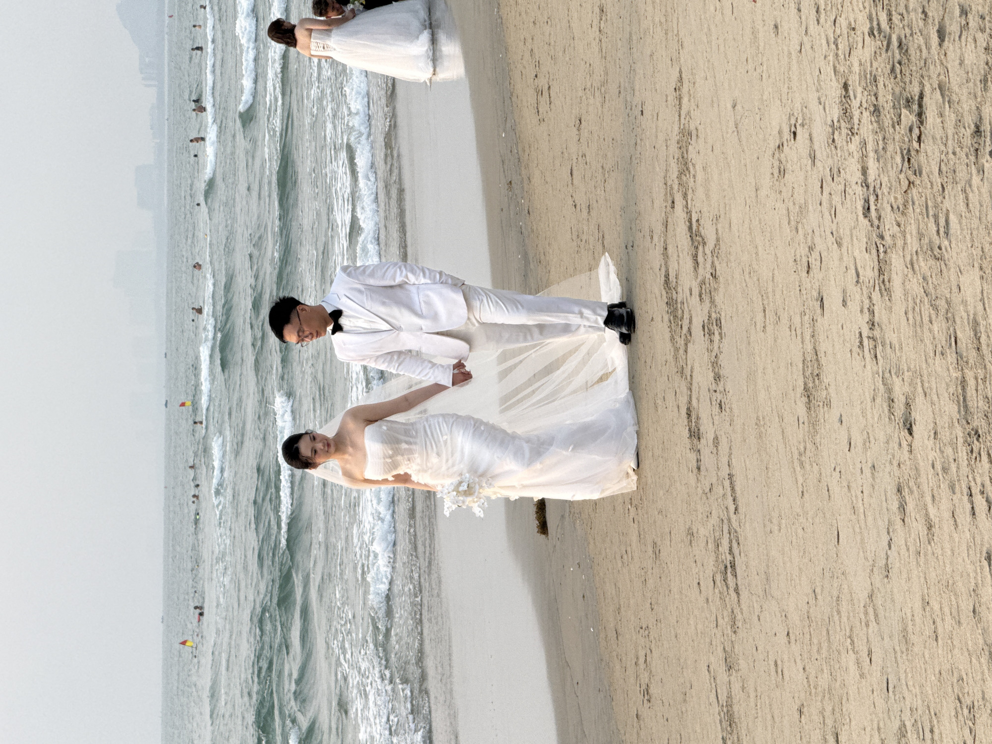 A couple is posing for a wedding photo on a beach. The groom is wearing a white suit and the bride is wearing a white wedding dress. The couple is holding hands and looking at each other. In the background, there is the ocean with waves and the city skyline of Đà Nẵng, Vietnam. There are other individuals at the beach further into the ocean. Another couple is positioned ahead of them in the same manner.