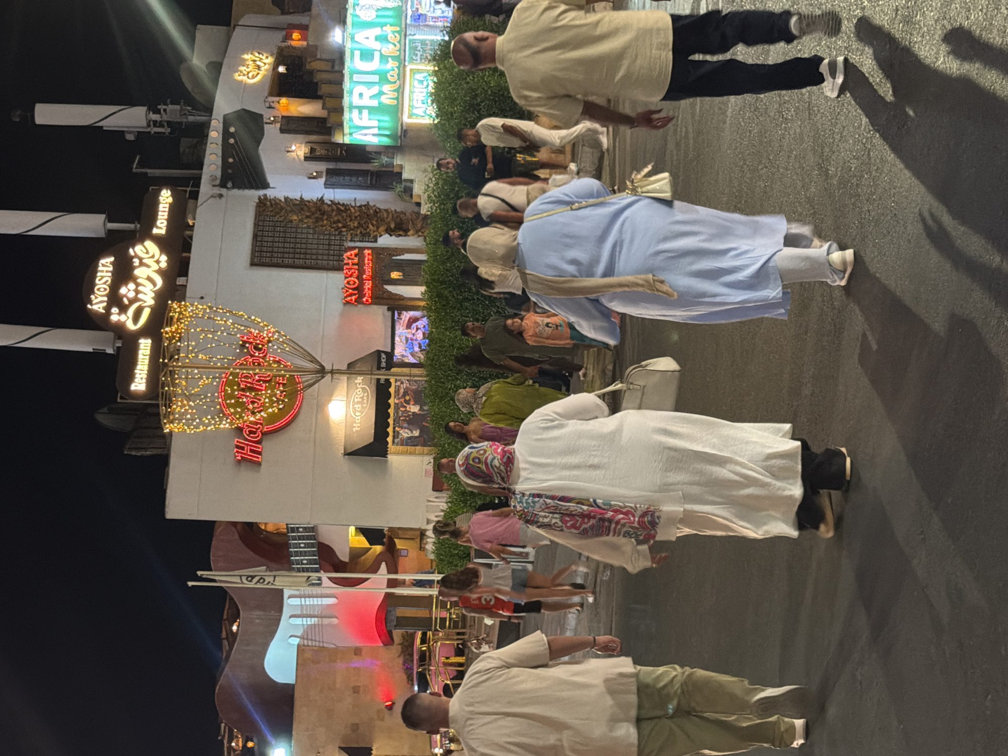 The image depicts a nighttime scene on a street in Sharm El Sheikh, Egypt. Several people are walking on the pavement. In the background, there are buildings with illuminated signs. One sign reads "AYOSHA Restaurant Lounge" in English and Arabic script. Another prominent sign displays "AFRICA Market" in bright green lettering. There are also decorative lights strung up, creating a festive atmosphere. The overall impression is a bustling commercial area at night, likely a tourist destination.