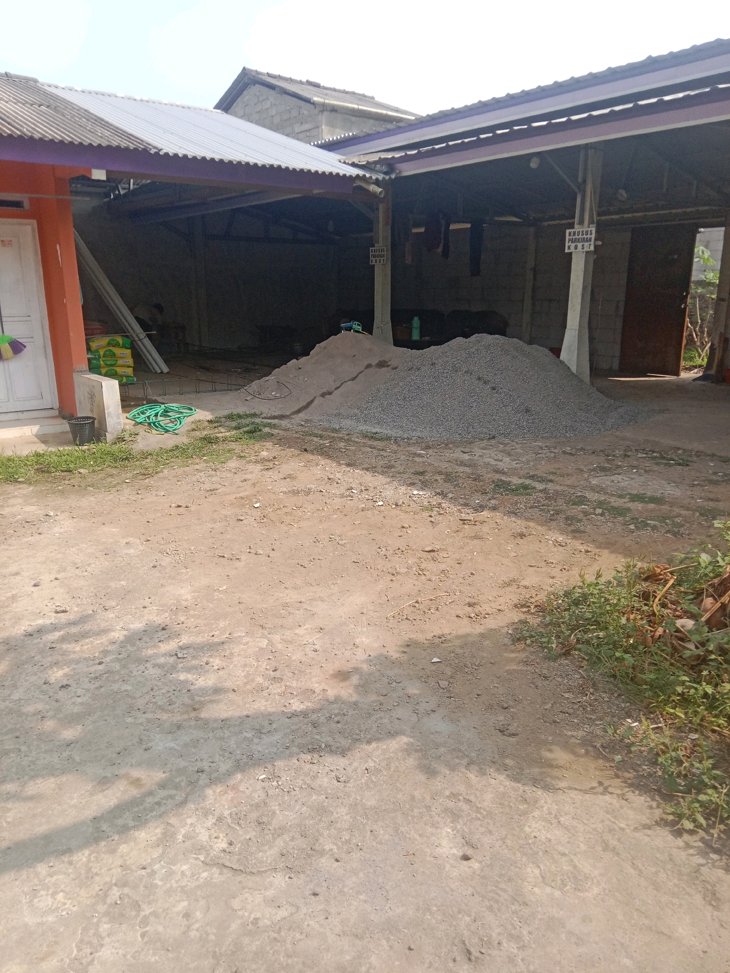 The image depicts an outdoor scene in the city of Leksono, Indonesia. In the foreground, a dirt and concrete ground is visible, with shadows cast by unseen objects. To the left, an orange building with a white door is partially visible, with a broom leaning against it and a black trash can. Adjacent to the building, a pile of green hoses is coiled on the ground.
In the middle ground, a large mound of grey gravel or small stones dominates the area. Behind the gravel pile, a covered outdoor structure with concrete pillars and a corrugated metal roof is present. Signs are attached to some of the pillars, one of which reads "KHUSUS PARKIR KOST". A few items of clothing hang from a line within the covered structure. In the background, the roofline of another building and a clear sky are visible. To the right of the covered structure, a dark wooden door is open, revealing a glimpse of the interior. The overall environment suggests a construction or storage area.