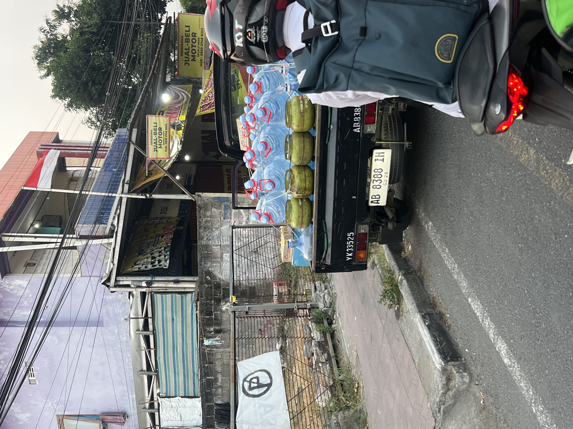 In the city of Sinduadi, Indonesia, a black pickup truck is parked by the roadside, its bed loaded with approximately 20 large plastic water jugs and four green propane tanks. The water jugs are clear, with red caps, and are arranged in multiple rows. The propane tanks are positioned in front of the water jugs. A white banner with a black circular logo is attached to a metal gate behind the truck. The truck has license plates that read "YK 33525" and "AB 8388 IH". In the foreground, a person on a motorcycle faces away from the camera, wearing a dark helmet with red accents and a dark teal backpack with a yellow circular patch. The scene appears to be in front of a building with a purple facade and several yellow signs advertising "MENERIMA JUAL-BELI MOTOR" (Accepts Buying-Selling Motorcycles). The sky is overcast, and there are many overhead power lines. The ground is asphalt with a concrete curb.
