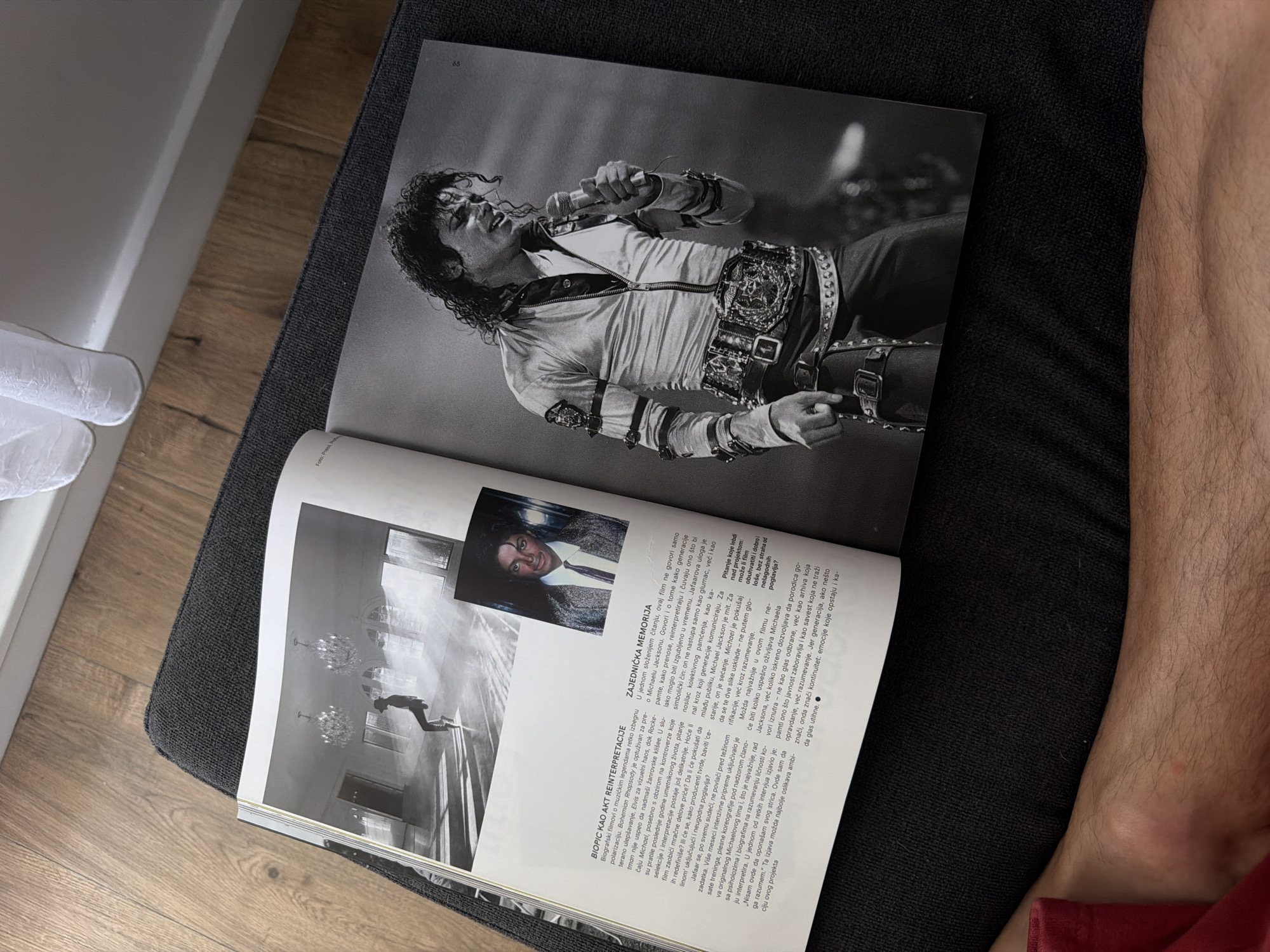 The image shows an open book with black and white photographs of Michael Jackson. The left page features a photograph of Jackson in a large, empty room with chandeliers, possibly a dance studio or a ballroom, with smoke or mist in the air. Beside this photograph, text is visible in a foreign language, likely Serbian, with a heading that appears to be about a biopic. The right page displays a full-page photograph of Michael Jackson performing on stage, holding a microphone and wearing a distinctive outfit with a large, ornate belt. Text is also present on this page. The book is placed on a dark gray upholstered surface, which could be a couch or a chair. Part of a person's bare leg, with dark hair, is visible in the lower left corner of the image, indicating someone is likely reading the book. The background includes a wooden floor and a white curtain. The scene is indoors. There is no information in the image or context to suggest a location in Palilula Urban Municipality, Serbia.
