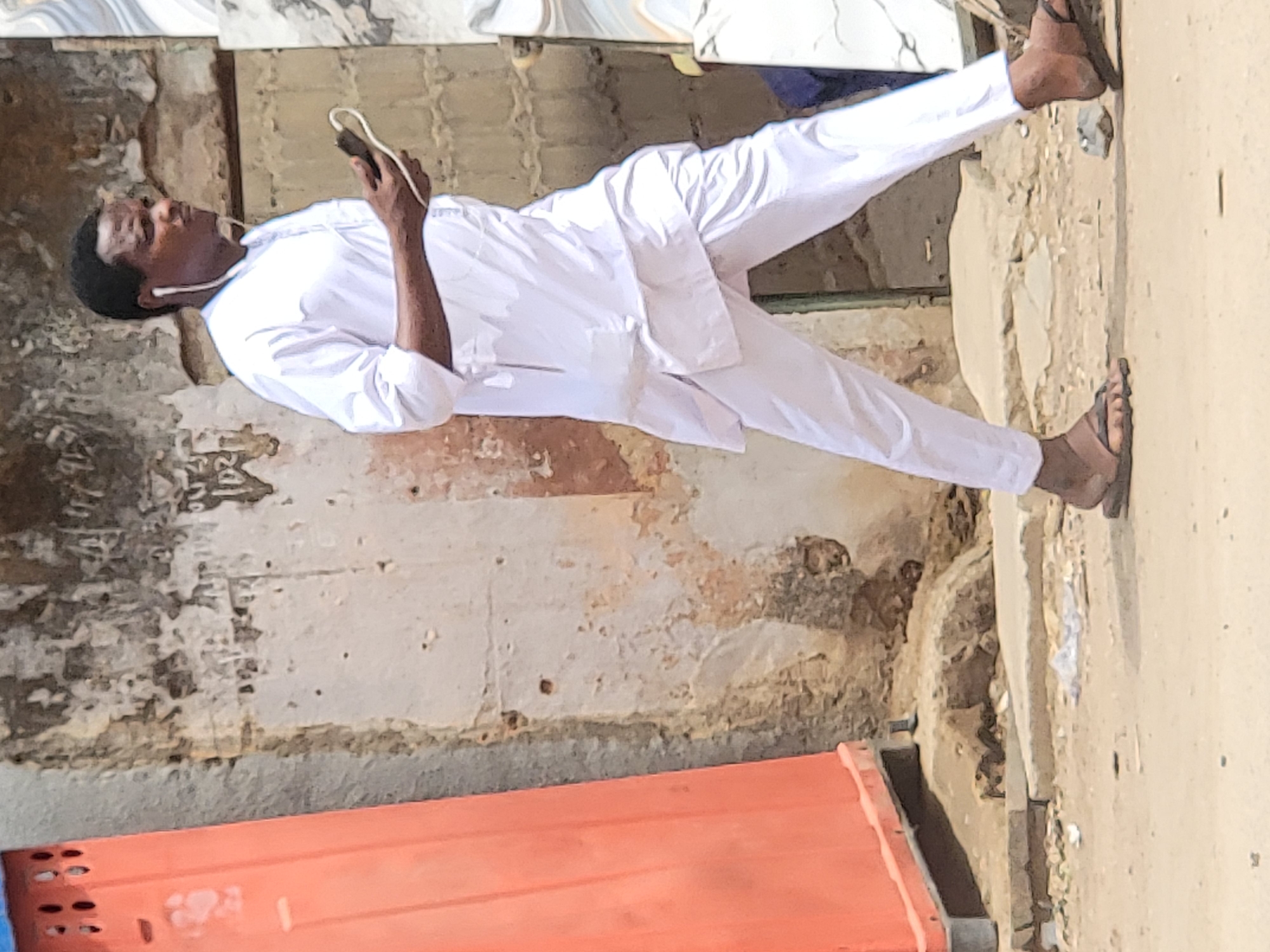 A man stands outdoors, dressed in a white traditional Nigerian outfit, consisting of a long-sleeved top and matching trousers. He wears dark sandals and has white wired earbuds in his ears, connected to a dark-colored smartphone he holds in his right hand. He appears to be focused on his phone, looking slightly upwards and to his left.
The setting is an outdoor area in what appears to be a residential or street environment in Maiburiji, Nigeria. The ground beneath him is sandy or dirt. Behind the man is a weathered wall, possibly part of an old building, with light-colored paint flaking off to reveal darker, underlying material. The overall impression of the wall is one of age and wear. To the bottom left of the frame, a bright orange object, possibly a container or part of a structure, is visible. The lighting suggests it is daytime with bright, clear weather. There is no visible text in the image. The man's expression is one of calm focus on his device.