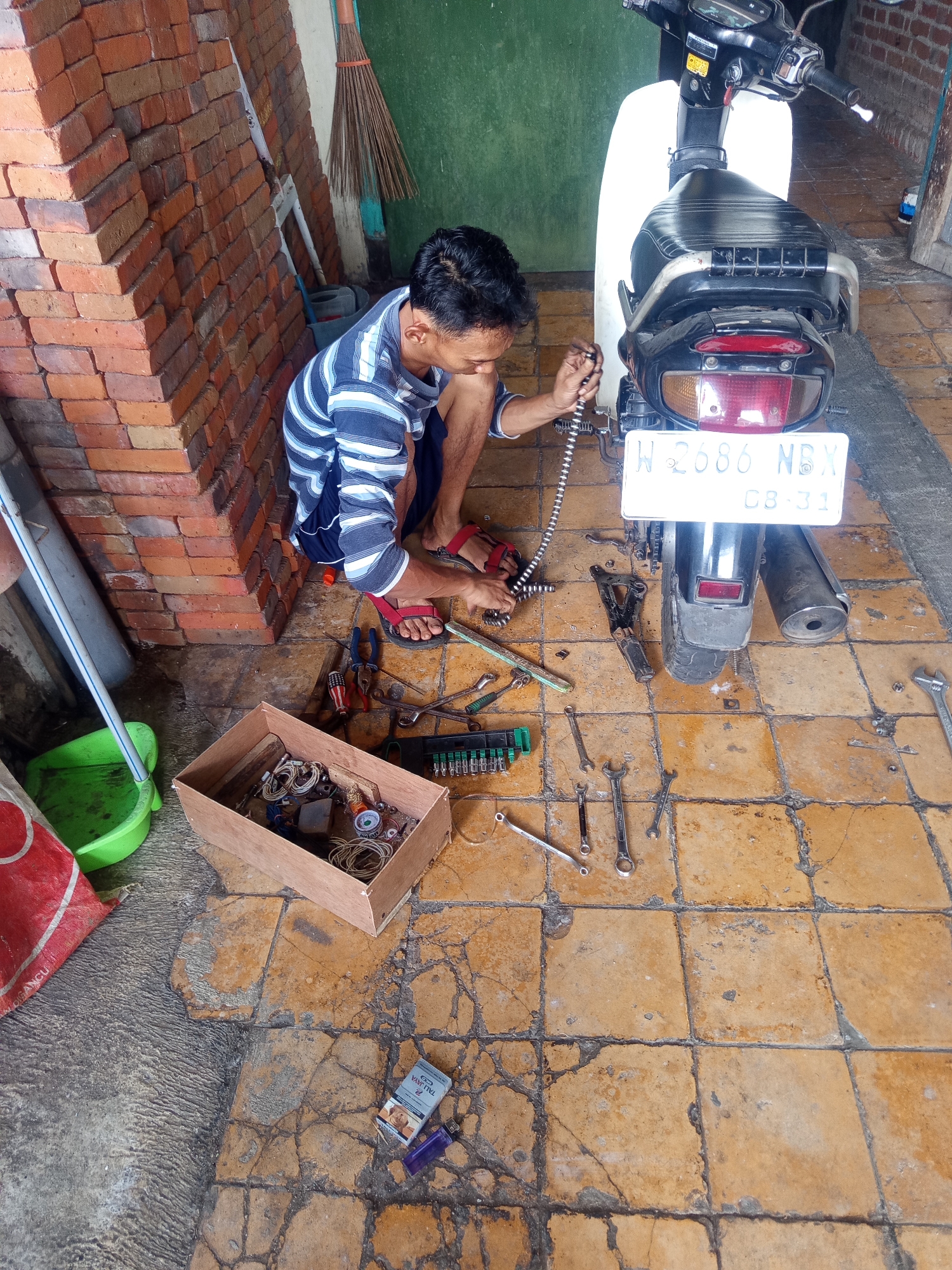 A male individual is crouched on a tiled floor in what appears to be an outdoor or semi-outdoor workshop area in Sumber Gedang, Indonesia, working on the rear section of a white and black motorcycle. He is wearing a blue and white striped t-shirt and dark shorts, with red sandals. The motorcycle has a white body, a black seat, and a license plate that reads "W 2686 NBX C8-31". Tools, including wrenches, pliers, and a screwdriver, are scattered on the floor around the motorcycle. A wooden box filled with various small items, possibly spare parts or hardware, is also present. To the left of the scene, a stack of red bricks is visible, along with a green bucket and a red bag. A broom with a wooden handle leans against a green wall in the background. A pack of cigarettes and a lighter are on the floor to the right, near the bottom of the frame.