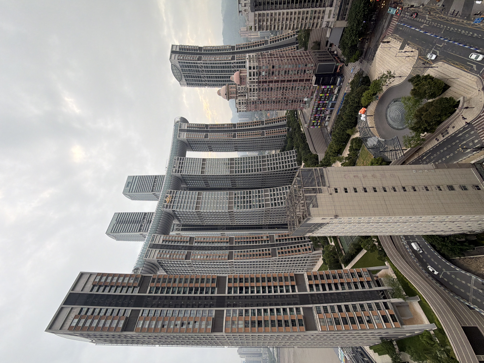 The image captures an aerial view of the Yuzhong District in Chongqing, China, featuring a striking modern architectural complex. Dominating the scene are several very tall, slender skyscrapers, many with curved or angled facades, clustered together. A distinctive feature is a skybridge that spans across the tops of some of these buildings, connecting them and forming a unique architectural element. The skybridge appears to be enclosed with a glass-like material and has a curved, segmented design.
Below the skyscrapers, a multi-layered road network winds through the urban landscape. Cars and at least one white van are visible on these roads, suggesting moderate traffic. A public square or plaza area is situated adjacent to the roads and some buildings, featuring a circular, amphitheater-like design with trees and paved walkways. A low-rise building with colorful signage is visible in the background, possibly a commercial establishment. In the foreground, another tall, light-colored building with a grid-like facade rises up, partially obscuring the view of the main architectural complex from this perspective.
The environment appears to be overcast, with a diffused light characteristic of a cloudy day. The overall impression is one of a dense, modern urban environment with ambitious and visually distinctive architecture. The presence of greenery, such as trees in the plaza and along the roadside, offers a contrast to the concrete and glass structures. The river can be glimpsed on the far left of the image, indicating the city's proximity to a major waterway.
