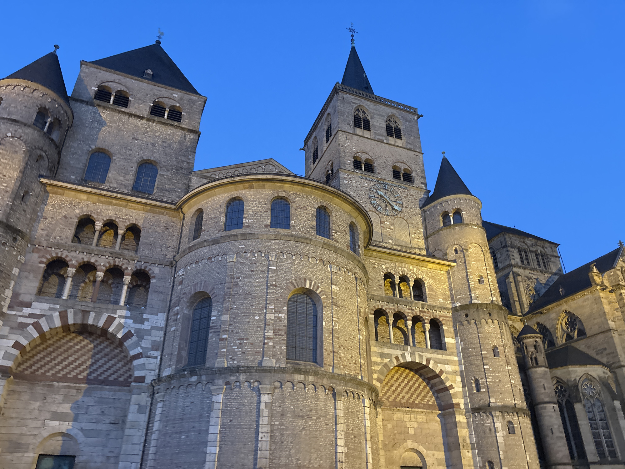 The image shows the Trier Cathedral, also known as the Dom St. Peter, in Trier, Germany, at dusk. The cathedral is a large Romanesque basilica with multiple towers, conical roofs, and intricate stone detailing. The sky is a deep blue, indicating the twilight hour. The stone facade of the cathedral is illuminated by artificial lights, highlighting its architectural features such as arched windows, decorative stonework, and a large clock face on one of the towers. There are no people visible in the image. The perspective is from a low angle, looking up at the imposing structure. The architecture is characterized by its massiveness, with heavy stonework and a mix of rounded and pointed arches. The overall impression is one of grandeur and historical significance.