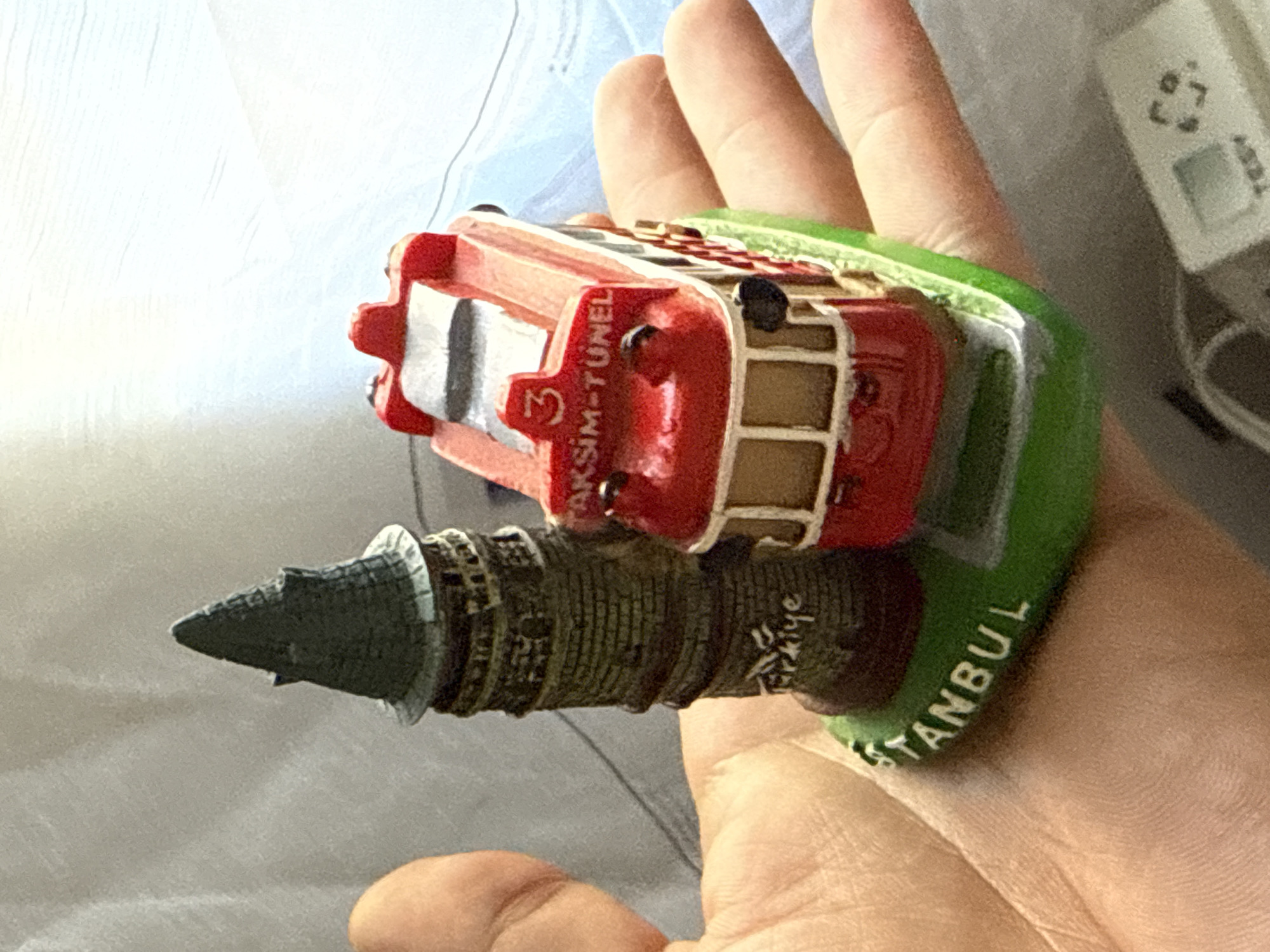 A hand holds a miniature souvenir depicting the Galata Tower and a red tram. The Galata Tower replica is dark green and textured to resemble brickwork, with a conical roof. The tram is bright red with white trim and the number "3" and "TAKSiM-TÜNEL" written on its front. The base of the souvenir is green, with "STANBUL" written in white letters. The background is out of focus and appears to be a light-colored fabric. The image does not show the city of Slavyani, Bulgaria.