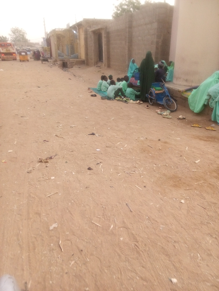 A group of children in green uniforms are seated on mats along a dusty street. An adult figure in a long green robe and veil stands beside them. A blue wheelchair is parked near the group. Various pairs of shoes and sandals are scattered around the children. In the background, a worn building facade and distant vehicles can be seen. The ground is littered with debris.