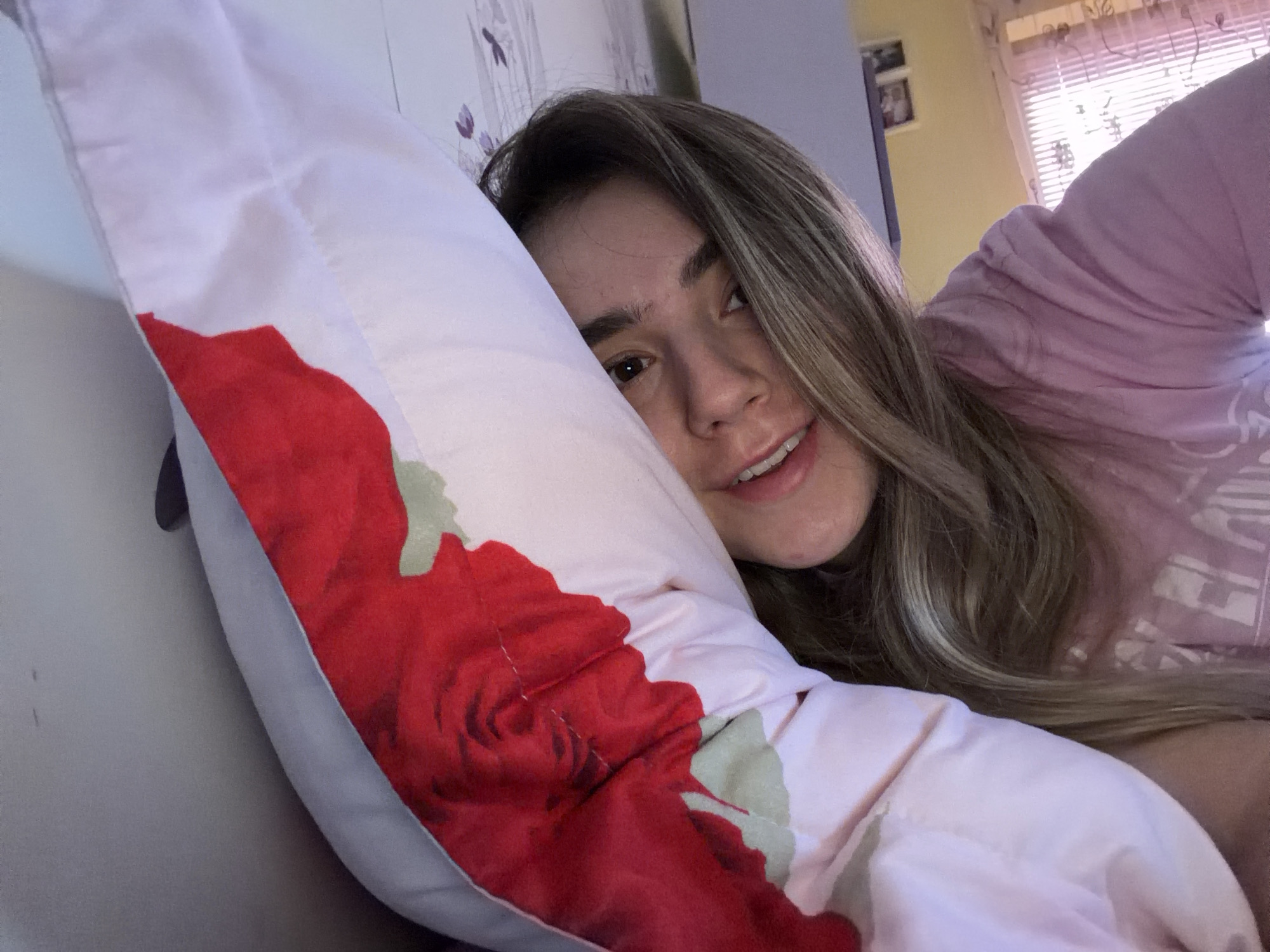 A woman is lying on a bed, partially covered by a pink and white pillow with a large red rose design. She is wearing a light pink top and has long, blonde hair with lighter highlights. The background includes a wall with a floral design, a window with blinds, and a few framed pictures. The location is an unknown city and state.