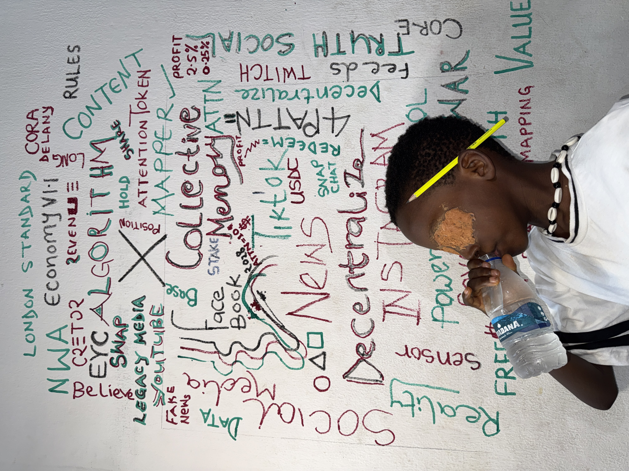 A young person with dark skin, a pencil behind their ear, and a substance smeared on their face is drinking from a water bottle. They are wearing a white shirt and a necklace with shells and beads. In the background, there is a wall covered in words and symbols in a mix of colors, including green, black, and red. The text includes words like "LONDON STANDARD", "ALGORITHM", "SOCIAL", "NEWS", "CONTENT", "ATTENTION", "TRUTH", "VALUE", and the names of social media platforms. The location is Ado, Nigeria.