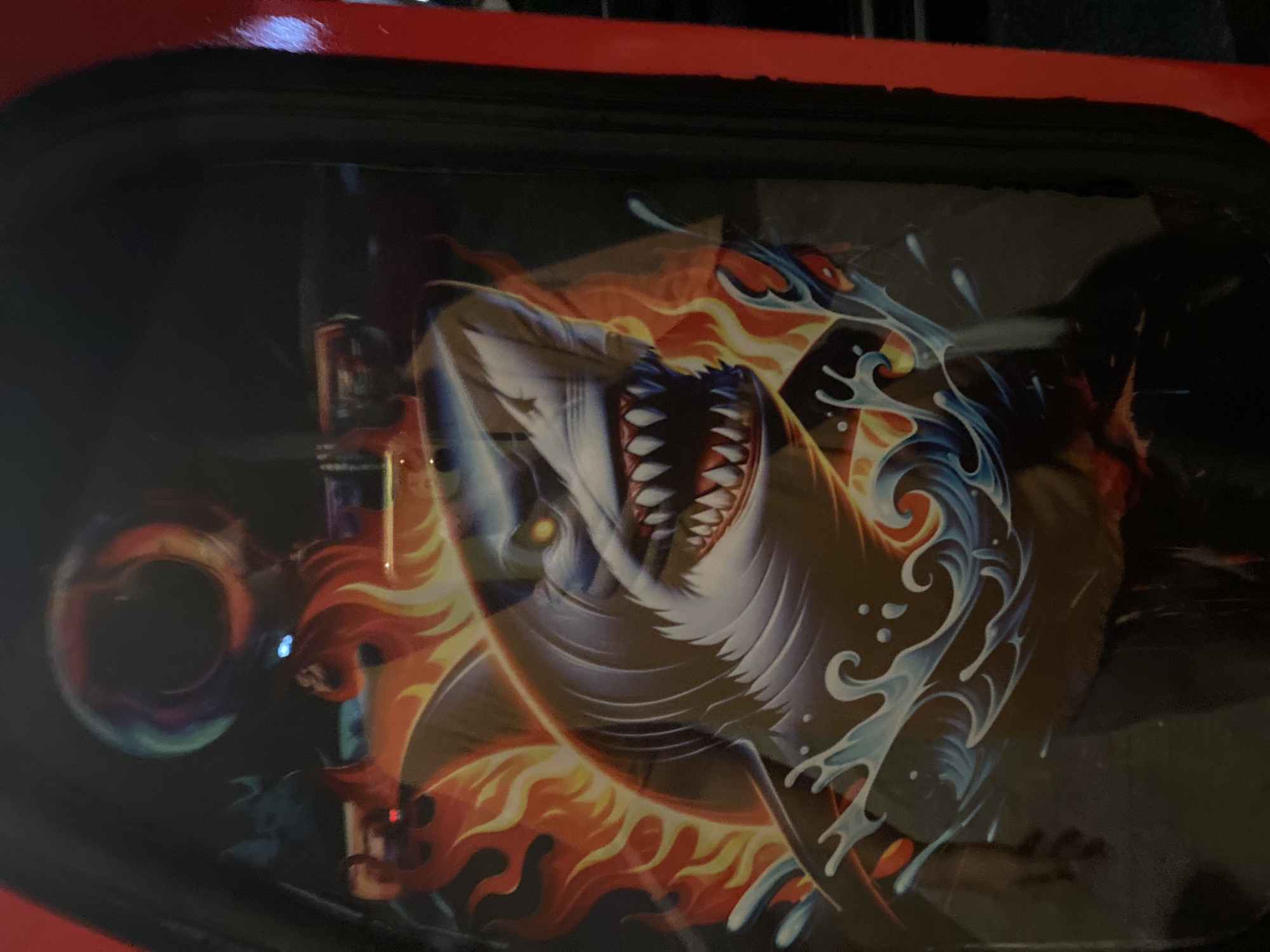 The image presents a vibrant graphic depiction of a shark, characterized by its grey body, prominent dorsal fin, sharp white teeth, and glowing yellow eyes, with its mouth agape in an aggressive posture. The shark is surrounded by stylized orange and red flames, indicating a sense of intensity or power, while below it, blue and white water splashes are rendered with a dynamic, illustrative style.
The graphic is contained within a rectangular panel, bordered by a dark frame, with a prominent vertical red strip framing the right edge. This suggests the graphic is part of a larger structure, such as an arcade game machine, a vending machine, or a decorative display case, observed from a slight angle.
The surface of the panel exhibits notable reflections. In the upper center, a circular, multicolored light reflection is visible. Further left, a subtle blue light source is reflected. A significant reflection in the lower-left quadrant clearly shows a human hand and forearm, along with a portion of a person's torso and legs, suggesting an individual is standing in front of the display. The reflected environment appears to be an indoor public space with ambient or low lighting, showing indistinct forms in the background.
This scene is observed within Davao City, Philippines.