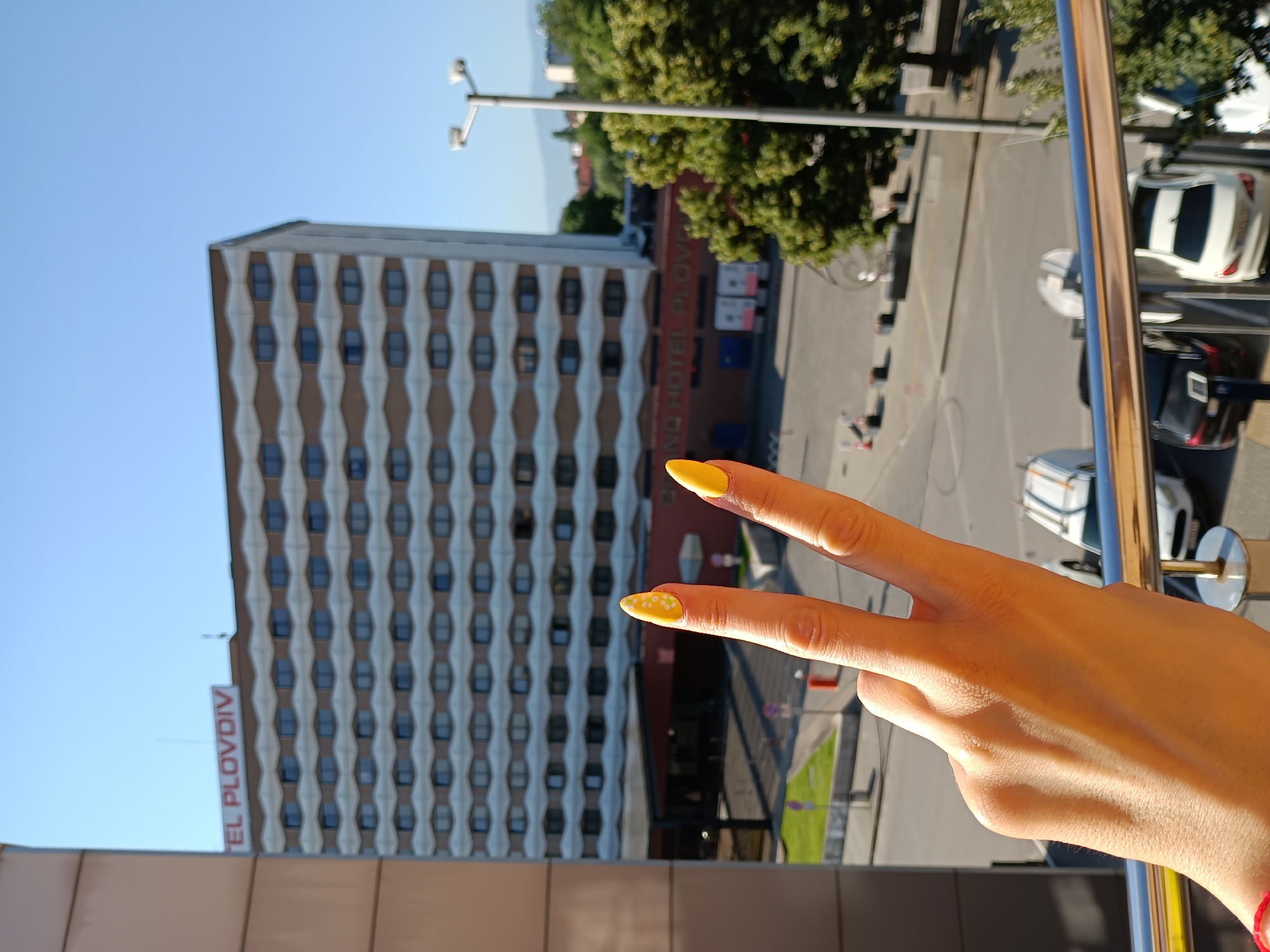 A person's hand, with bright yellow fingernails decorated with small white polka dots, is held up in a peace sign gesture, obscuring the lower left portion of the frame. Behind the hand, a large hotel building with a distinctive diamond-patterned facade dominates the background under a clear blue sky. A red sign with white lettering reads "HOTEL PLOVDIV" across the top of the hotel's entrance area. The street level below the hotel shows parked cars, including a white van and a white sedan, and some trees. The image is taken from a slightly elevated perspective, possibly from a balcony or window, indicated by a metal railing in the foreground. The scene suggests a daytime setting in the city of Plovdiv, Bulgaria.