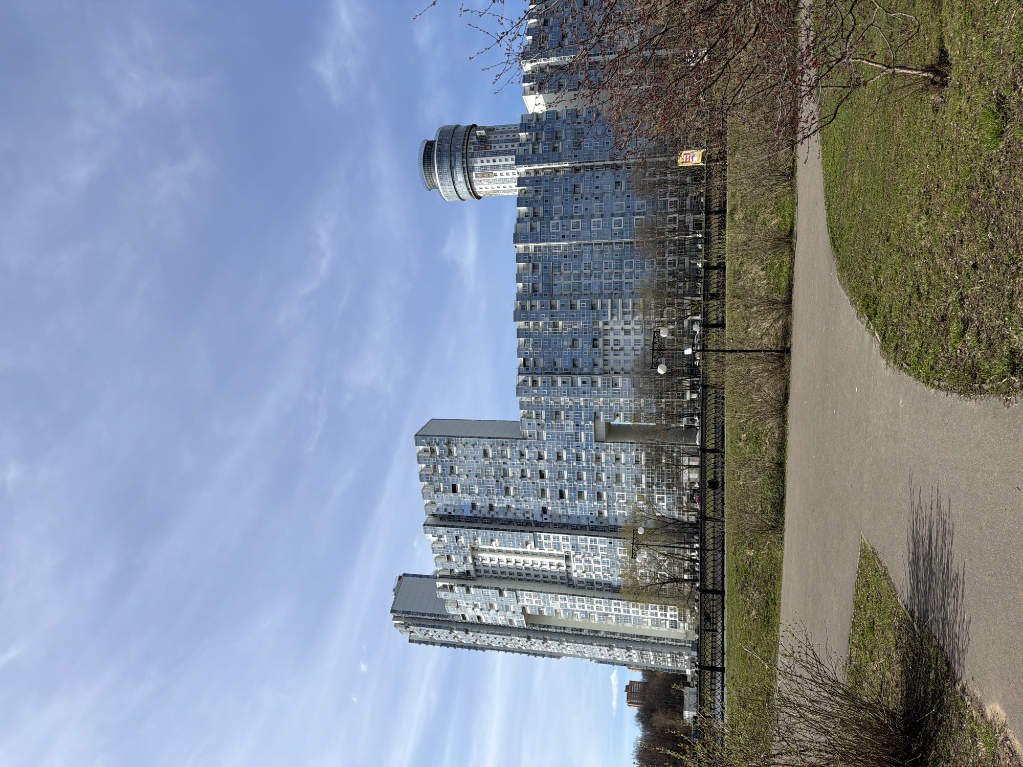 The image shows a modern apartment complex in Khimki, Russia, on a bright, sunny day. The buildings are tall and have a unique architectural design, with numerous windows and a cylindrical structure atop one of the towers. A paved pathway runs alongside the complex, separated by a low, black fence. The pathway is flanked by grassy areas and some trees. No people are visible in the frame.