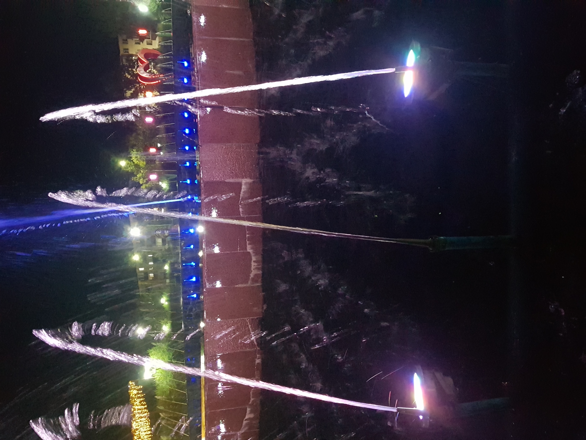 The image captures a night scene of a fountain with illuminated water jets in Kurchatov, Kazakhstan. The fountain is the primary subject, with several water streams arcing upwards, lit by colorful lights. The base of the fountain is a dark pool of water, reflecting the lights and the water spray. Behind the fountain, a multi-tiered structure, possibly part of a building or embankment, is visible with reddish-brown textured surfaces. Further in the background, there are trees, streetlights, and a building with illuminated windows. Decorative lights, appearing as yellow string lights and red tulip-shaped lights, are also present in the background, along with a red heart-shaped sculpture. The overall atmosphere is festive and illuminated, typical of an outdoor evening attraction. There are no people visible in the image.