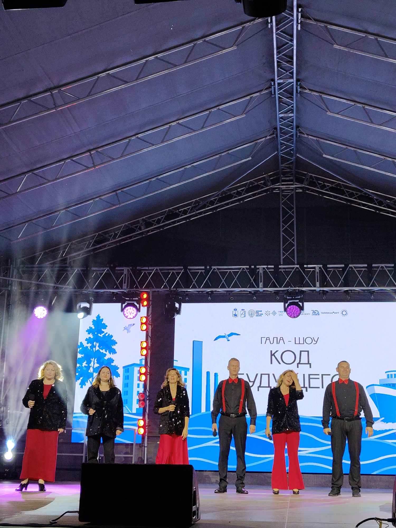 Six performers, three women and three men, are on a brightly lit stage in front of a large screen displaying graphics. The women are dressed in black sequined jackets and long red skirts, or red pants. The men are dressed in black shirts with red suspenders and red bowties, and black pants. The stage is equipped with lighting and sound equipment, and a large black speaker is positioned in the foreground. The screen behind the performers shows a blue graphic with a tree and a bird. The overall scene appears to be a live performance event.