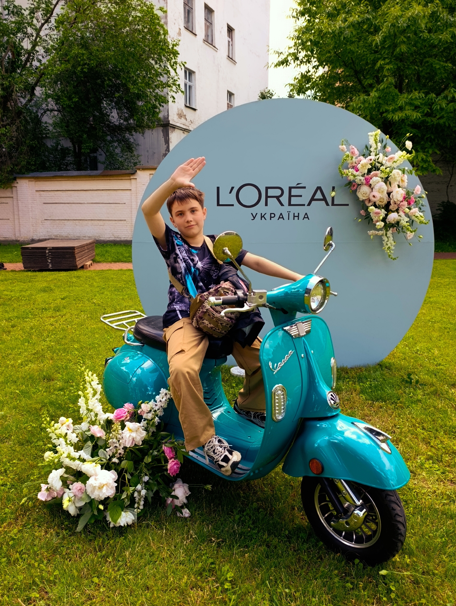 A young boy with short brown hair, wearing a dark t-shirt with a graphic print and khaki pants, is sitting on a teal scooter. He has his right arm raised in a wave. The scooter is decorated with a bouquet of pink and white flowers at its front. Behind the scooter is a large, light blue circular backdrop with the L'Oréal logo and "Україна" (Ukraine) written in black text. The backdrop is also adorned with a floral arrangement. The scene is set outdoors on a grassy area, with a white brick wall and trees visible in the background, suggesting an urban environment like Kyiv, Ukraine. The overall lighting indicates daylight.