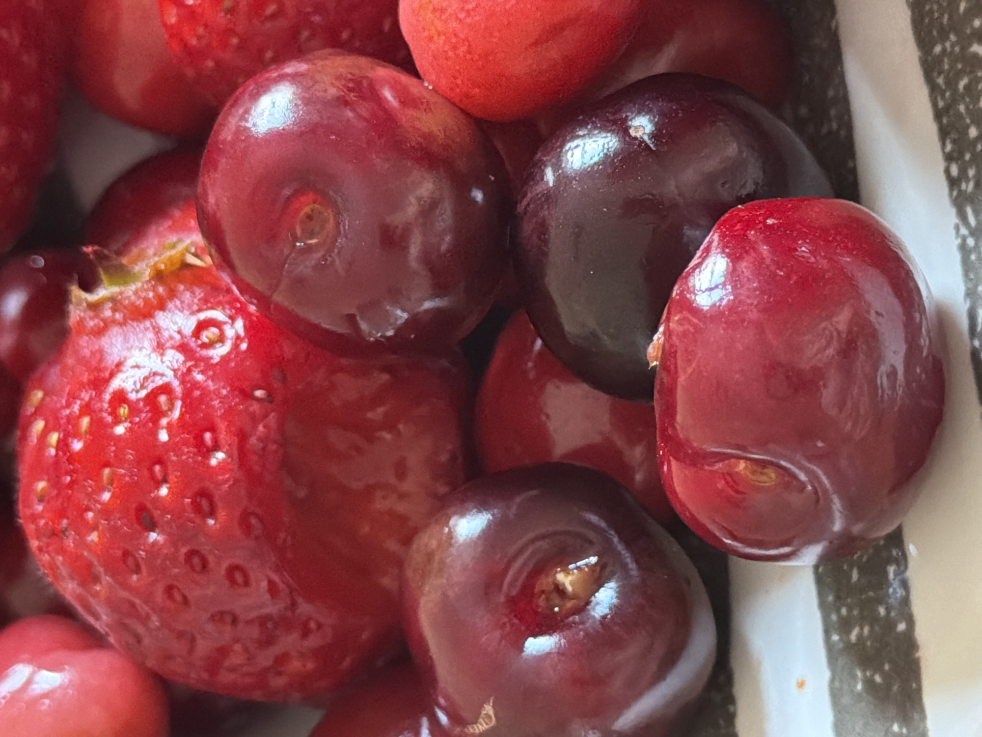 A close-up shot shows a collection of red and dark red fruits, specifically strawberries and cherries, clustered together in what appears to be a container. The strawberries are bright red and textured with small seeds. The cherries are round, glossy, and range in color from a lighter red to a deep, almost black, hue. The background is indistinct, with glimpses of a black and white striped pattern suggestive of the container's design. There is no indication of the location in Catania, Italy. No people, actions, or interactions are visible in the image. The focus is solely on the fruits and their immediate surroundings.