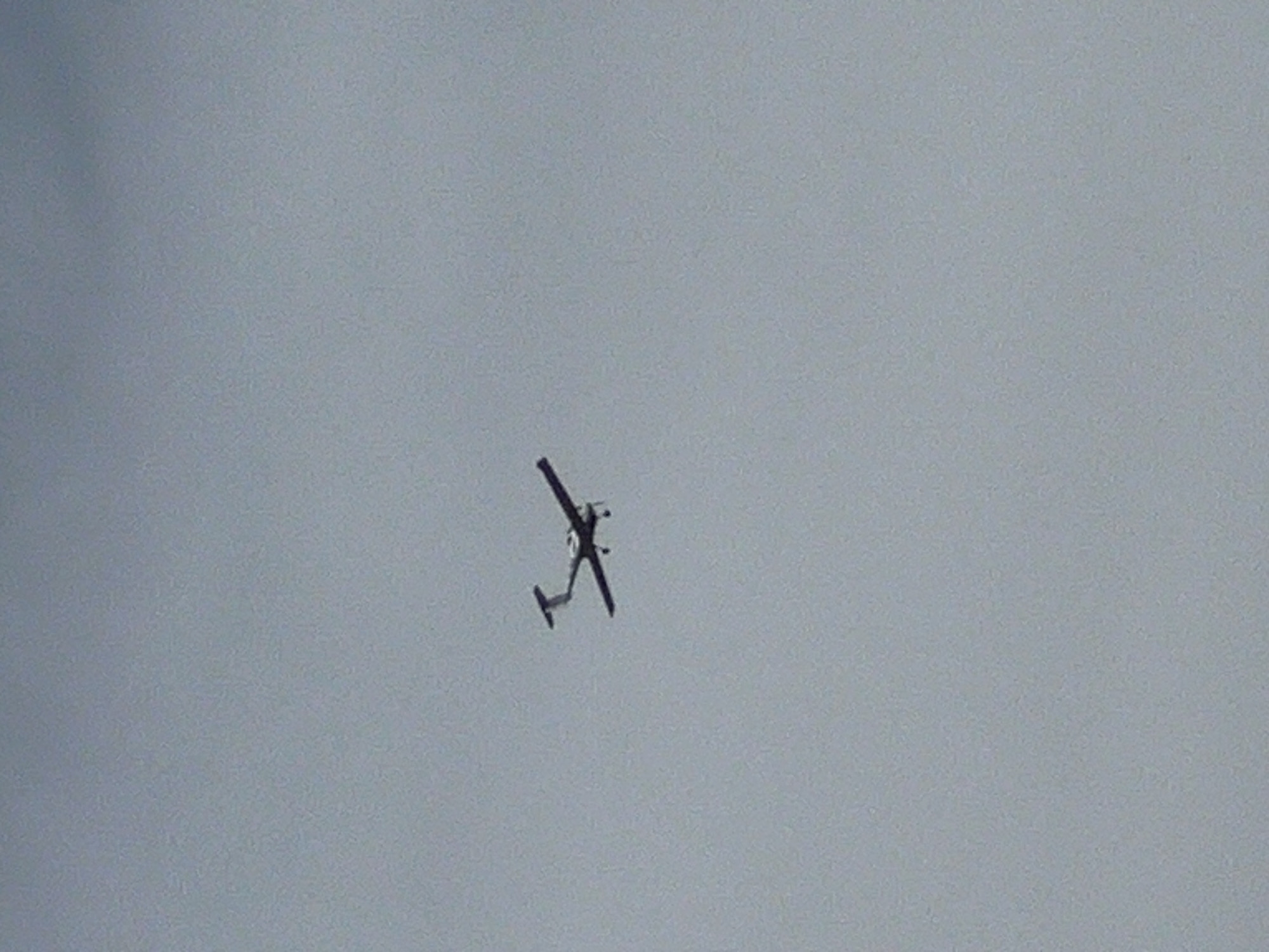 A small, single-engine airplane is captured in mid-flight against a uniformly overcast sky. The aircraft appears to be a low-wing monoplane with a tail rudder. Its body and wings are silhouetted against the pale sky. The image is a medium shot, with the airplane positioned slightly off-center to the right. The background is entirely composed of the sky, indicating an aerial or high-angle perspective. The image does not depict any elements that would suggest a specific location within the city of Lublin, Poland, or any urban environment.