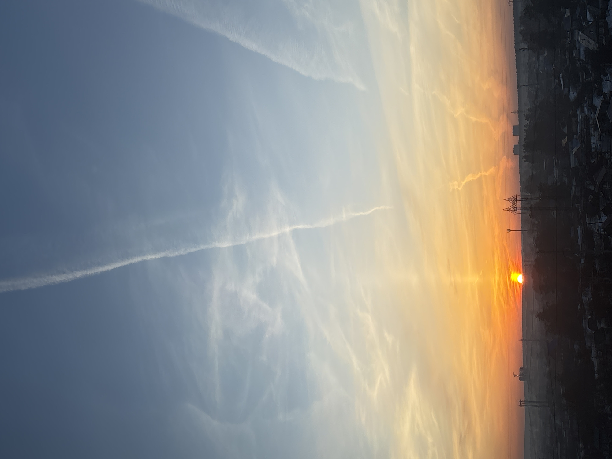 The image depicts a sunrise or sunset over Chaikovsky, Russia. The sky is divided into two distinct sections: a light blue expanse to the left, marked with contrails and wispy clouds, and an orange-yellow section to the right, closer to the horizon, where the sun is visible as a bright orange-red disk. The horizon line is cluttered with the silhouettes of buildings, trees, and electrical transmission towers, suggesting a cityscape. The light gradient indicates the sun is either rising or setting behind this urban landscape.