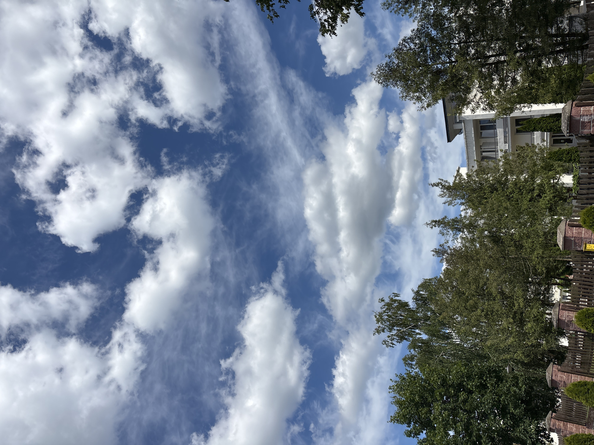 The image depicts a scenic view of a blue sky with scattered white clouds. The clouds are a mix of wispy cirrus formations and larger cumulus clouds, suggesting a dynamic weather pattern. The sky takes up a significant portion of the frame, creating a sense of vastness.
In the lower right portion of the image, trees with lush green foliage are visible, bordering a wooden fence. Behind the trees, a modern building with white walls and multiple windows can be seen. The architecture suggests a residential or possibly commercial property. The trees are casting shadows, and the sunlight filtering through the leaves indicates it is likely daytime, possibly late morning or early afternoon.
The overall mood of the image is peaceful and natural, with the bright sky and greenery creating a pleasant atmosphere. The location context provided is "Парэчча, Belarus," which places the scene in a specific geographical area. There is no visible text in the image. The composition is vertical, with the sky dominating the upper left and the trees and building occupying the right side.