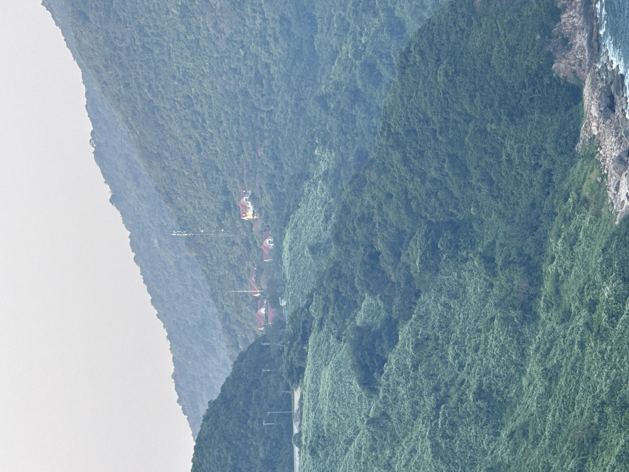 A landscape view of a mountainous area, potentially in the city of Đà Nẵng, Vietnam. The mountains are covered in dense green vegetation. Some buildings with red roofs can be seen on one of the mountain slopes. A road runs along the base of the mountain. The sky is overcast. Rocks and water are on the edge of the picture.