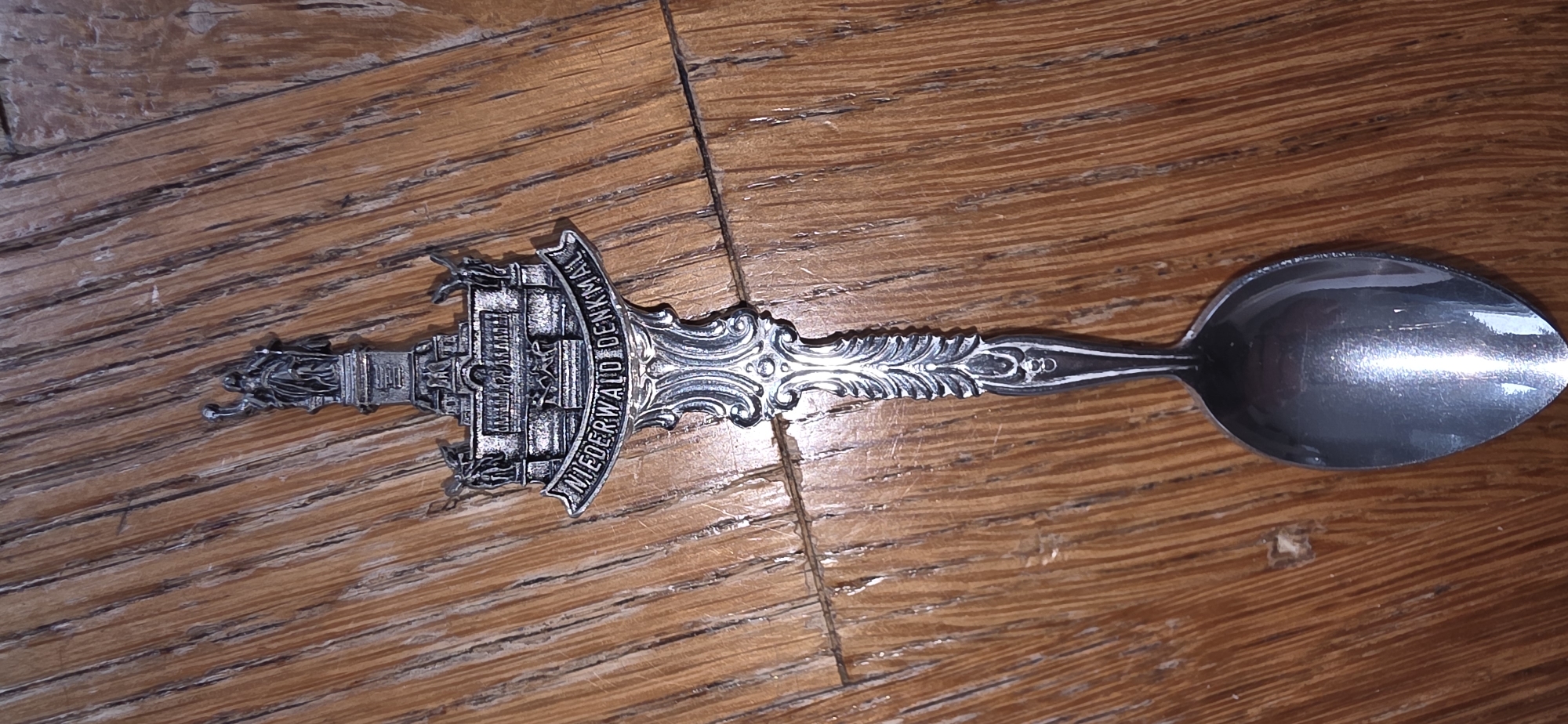 A detailed, ornate silver spoon lies on a wooden surface. The spoon's handle is decorated with intricate scrollwork and culminates in a detailed depiction of the Niederwald Monument. This monument features a prominent statue of Germania at the top, with smaller statues and architectural elements below, including the inscription "NIEDERWALD DENKMAL" across a banner. The wooden surface exhibits a prominent grain pattern and appears to be a table or floor. The image is a top-down, close-up shot, with the spoon centrally placed. The Niederwald Monument is located in Germany, not Marum, Netherlands.