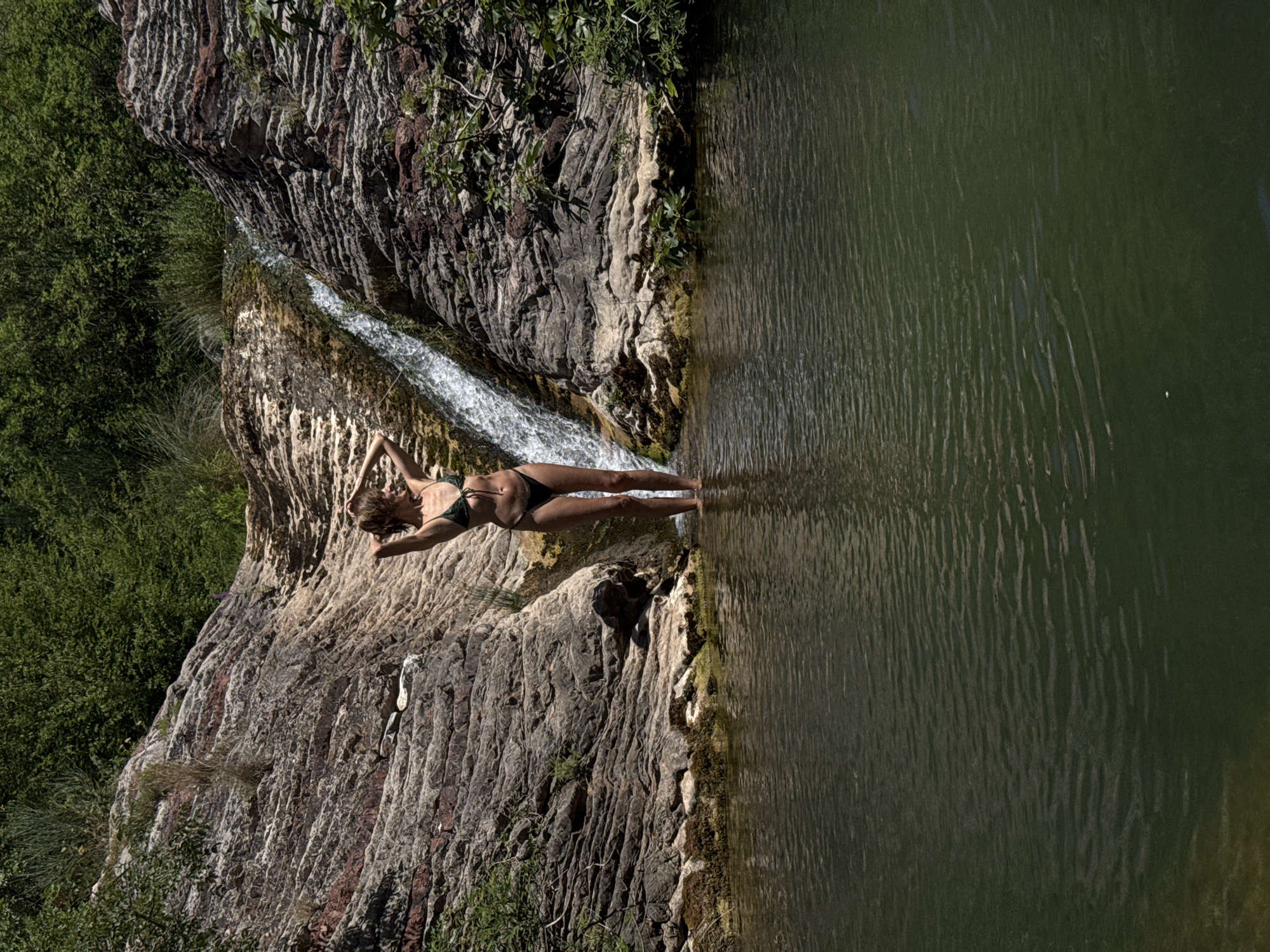 A woman in a dark green bikini stands in shallow water at the base of a small waterfall cascading over layered rock formations. She has her hands behind her head, and her body is slightly angled towards the viewer. The water in the foreground is dark and rippled, reflecting the surrounding greenery. Lush vegetation covers the top of the rock formations and spills down the sides. The overall environment suggests a natural, secluded outdoor setting. The location is identified as Rafailovići, Montenegro.