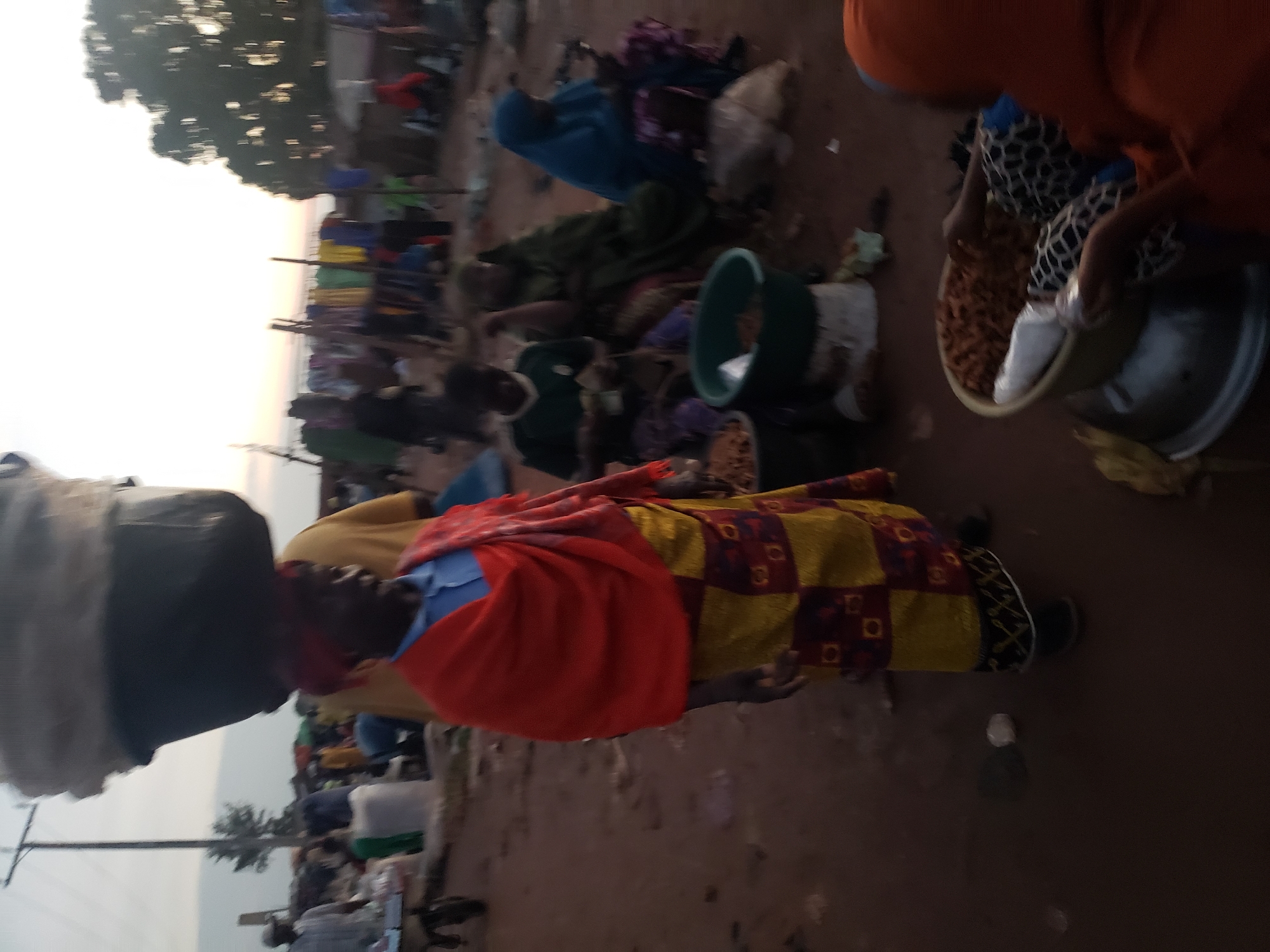 In the city of Kakuri Gwari, Nigeria, a busy marketplace scene unfolds at dusk. A woman in the foreground, with a vibrant red cloth draped over her shoulders and a yellow and red patterned skirt, balances a large, dark container of goods on her head. She wears a blue collared shirt beneath the draped cloth. Behind her, other market-goers are visible. To the left, a man in a light-colored shirt pushes a cart laden with goods. Further back, several people are seated, engaged in transactions or conversation. A woman in a green headscarf and dress sits in the center, and to her right, another woman in a blue hijab is looking down. Clothing hangs on display in the background, suggesting a vibrant commercial hub. The ground is a reddish-brown dirt, characteristic of the region. The overall atmosphere is one of lively activity as the day winds down.