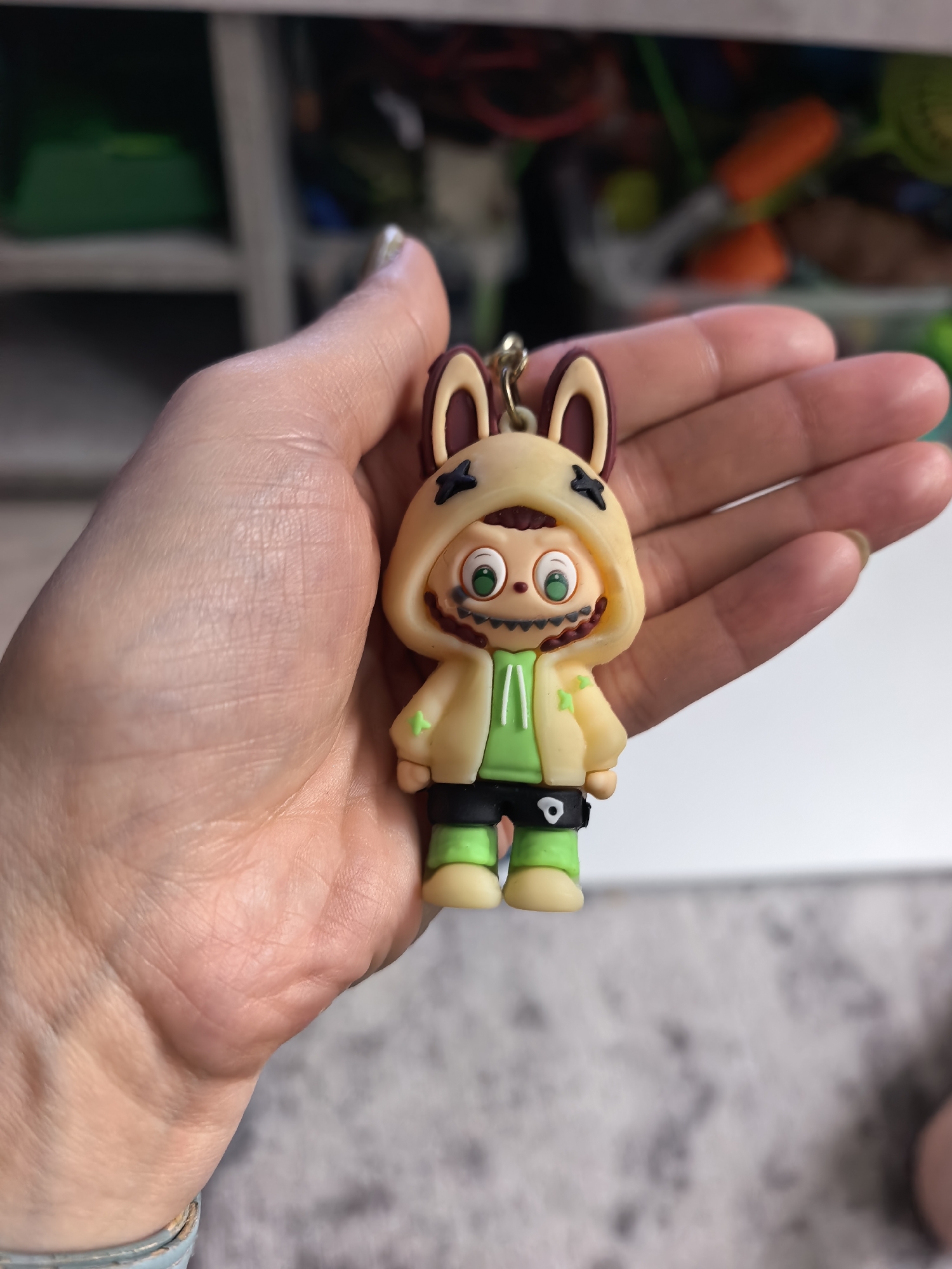 A hand holds a small, detailed keychain figure of a cartoon character with bunny ears. The figure wears a yellow hooded jacket with green accents and black shorts, with green shoes. The background shows shelves and various objects, suggesting an indoor setting like a store or a room. The location is not specified in the image or its context.
