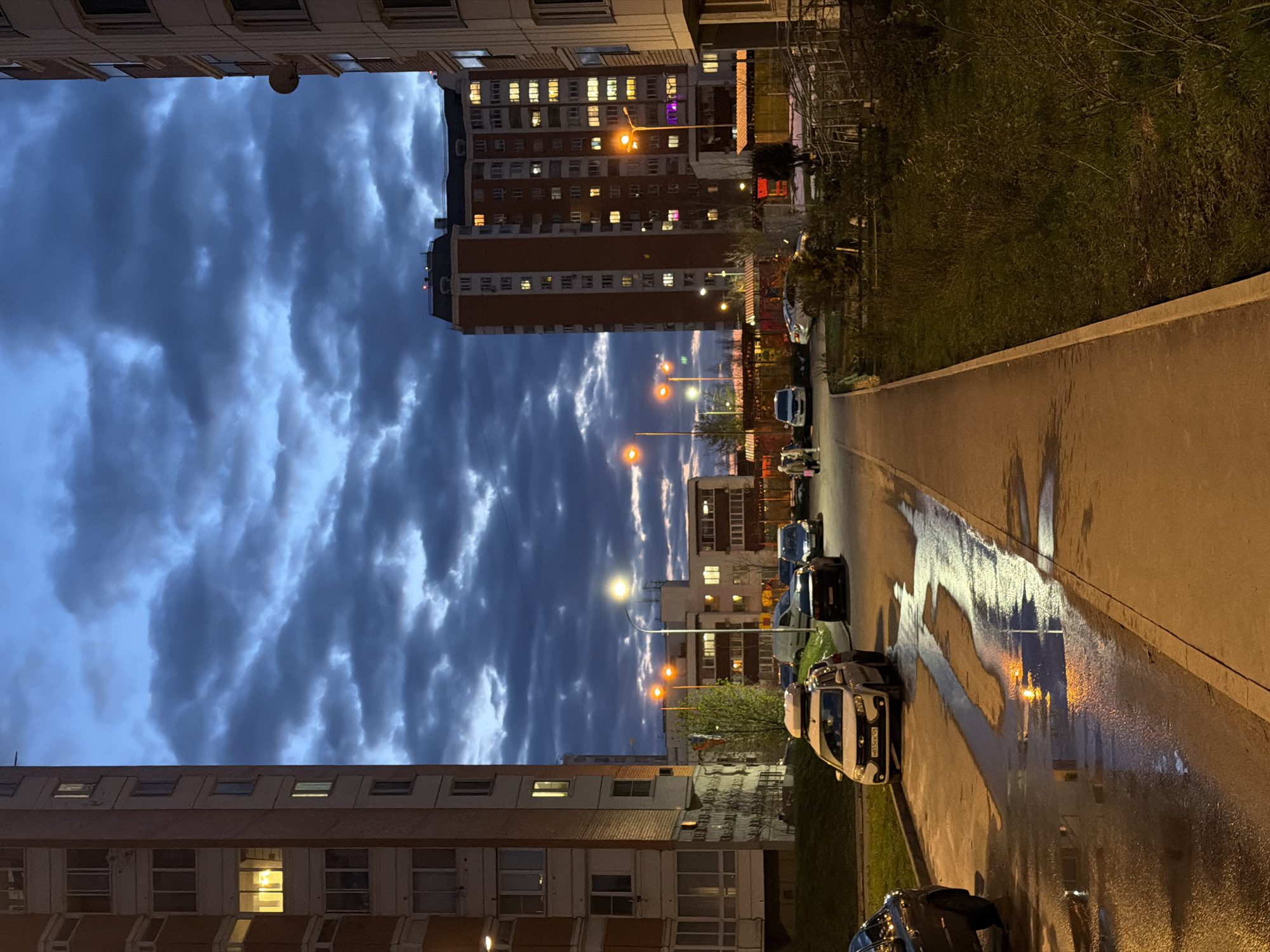 This media file captures an urban residential scene at dusk or early night, likely in Brekhovo, Russia. The sky is dominated by thick, dark blue-grey clouds, suggesting a recent rain shower or an overcast evening.
The scene features several multi-story apartment buildings. On the left side, a building with many windows, some illuminated, frames the view. In the middle ground, a prominent, tall building with reddish-brown facades stands out, its numerous windows casting warm yellow light onto the darkening surroundings. Further down the street, other residential structures are visible.
A paved road or pathway, slick with moisture from recent rain, runs along the right side of the image. The wet surface creates strong reflections, particularly from the warm orange glow of several streetlights that line the path. These reflections are elongated and distorted, adding a dynamic element to the damp ground.
Multiple cars are parked along the side of the road. One white car in the foreground prominently displays a Russian license plate: "O585CE 190". Scattered vegetation, including trees and bushes, can be seen along the road and between the buildings, appearing dark against the dim light. There are no clearly identifiable individuals, but some indistinct figures are visible further down the street, possibly pedestrians.
The overall mood is serene and quiet, characteristic of a residential area winding down for the evening, with the interplay of artificial lights, wet surfaces, and cloudy skies creating a subdued yet engaging atmosphere.
