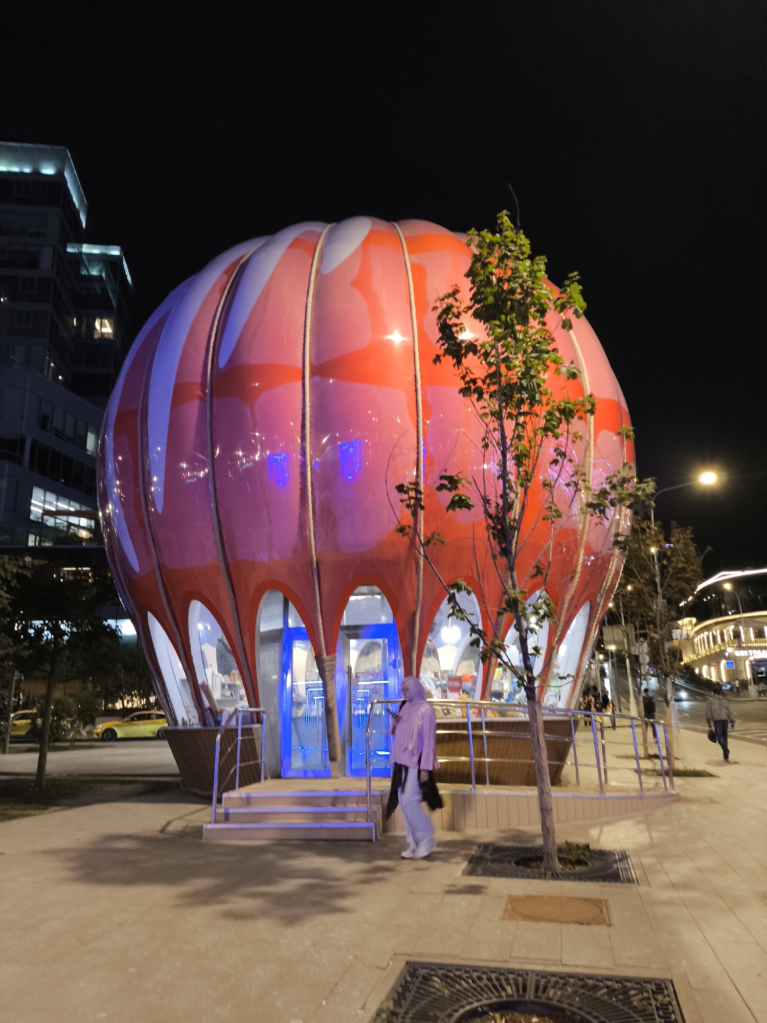 A night time, eye-level, medium shot shows a uniquely shaped red and white building resembling a hot air balloon. The structure is illuminated, with blue lights around its entrance and a warm glow from within. A young woman wearing a light purple hijab and matching pants stands at the entrance, facing the building. She is carrying a black bag over her shoulder. A small, bare-branched tree stands in front of the balloon-shaped structure, casting a shadow on the concrete ground. In the background, there are tall buildings with illuminated windows, a street lamp, and a few yellow cars parked on the side of the road. The scene is set in a city at night.