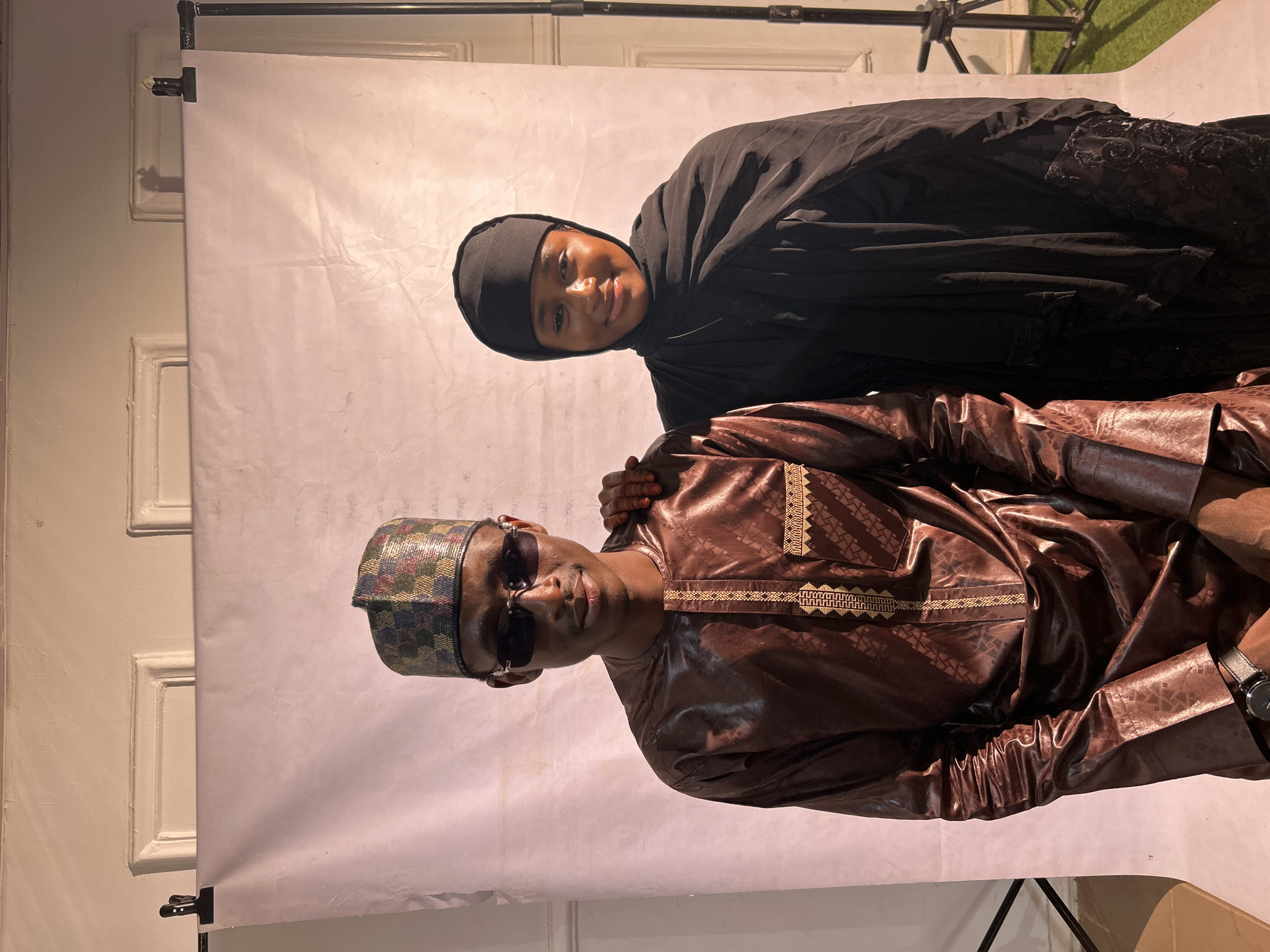 The image depicts a man and a woman posing in front of a white backdrop. The man is wearing a brown patterned tunic, sunglasses, and a cap with a multicolored pattern. He has a watch on his left wrist. The woman is wearing a black hijab. Her left hand is placed on the man's right shoulder. The backdrop is mounted on a stand, with a door visible on the left side of the frame and green material visible on the top right. The image was likely taken in a studio setting, given the backdrop and lighting. The location of the photo in Jos, Nigeria cannot be confirmed.