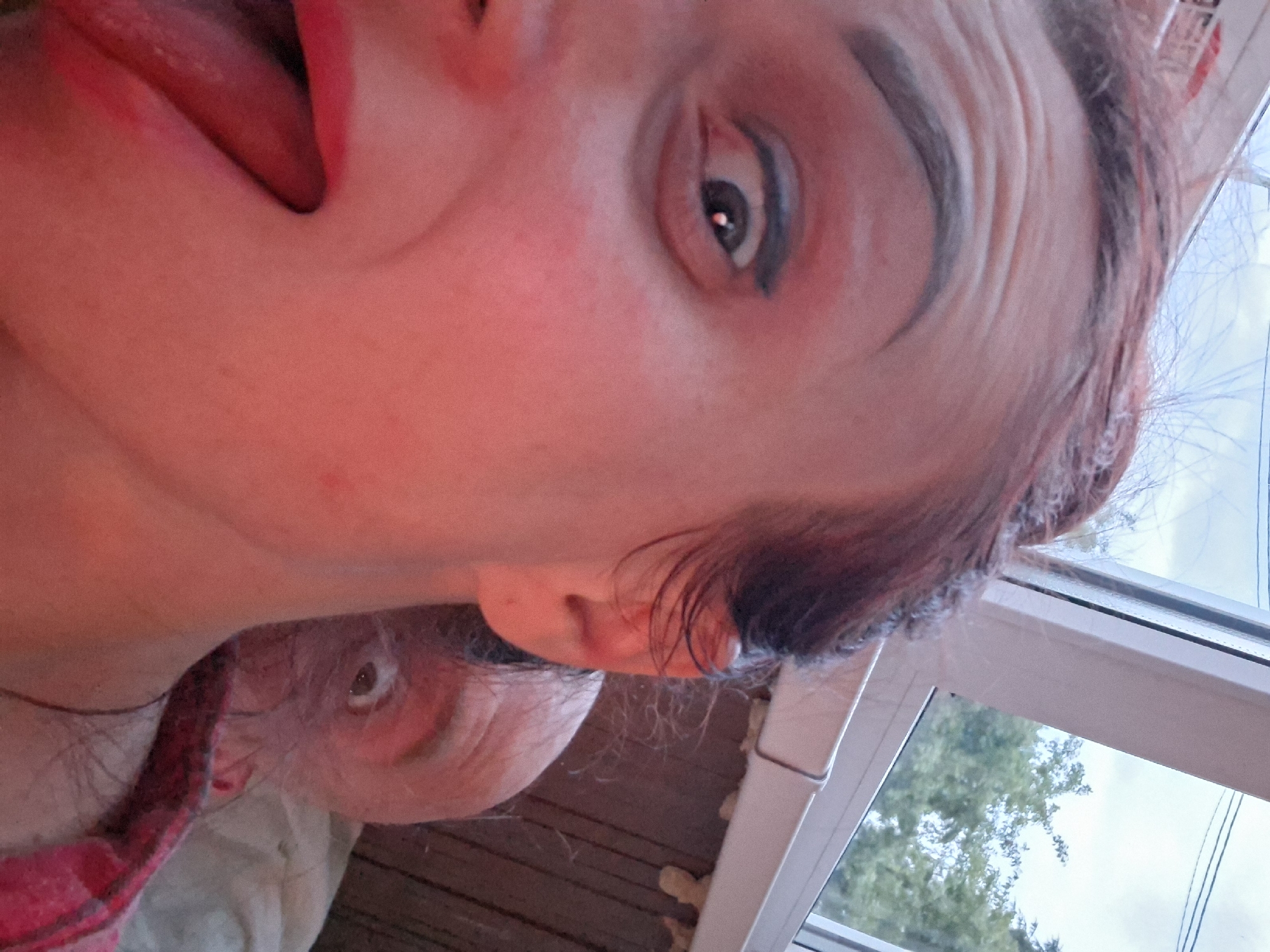A close-up, low-angle selfie shows a woman's face filling the left side of the frame, with her mouth open and tongue sticking out. Her eyes are wide and looking directly at the camera, with dark eyeliner applied. Behind her, partially obscured, a man's face with wide eyes and an open mouth is visible. They appear to be indoors, near a window with a view of trees and sky. The lighting is warm and reddish, possibly from artificial light. The background shows wooden paneling and insulation. The woman has dark hair pulled back, and is wearing a red and black plaid shirt. The man is wearing a light-colored garment. The context suggests a casual, possibly playful moment captured at a residence in Забалоцкі сельскі Савет, Belarus.