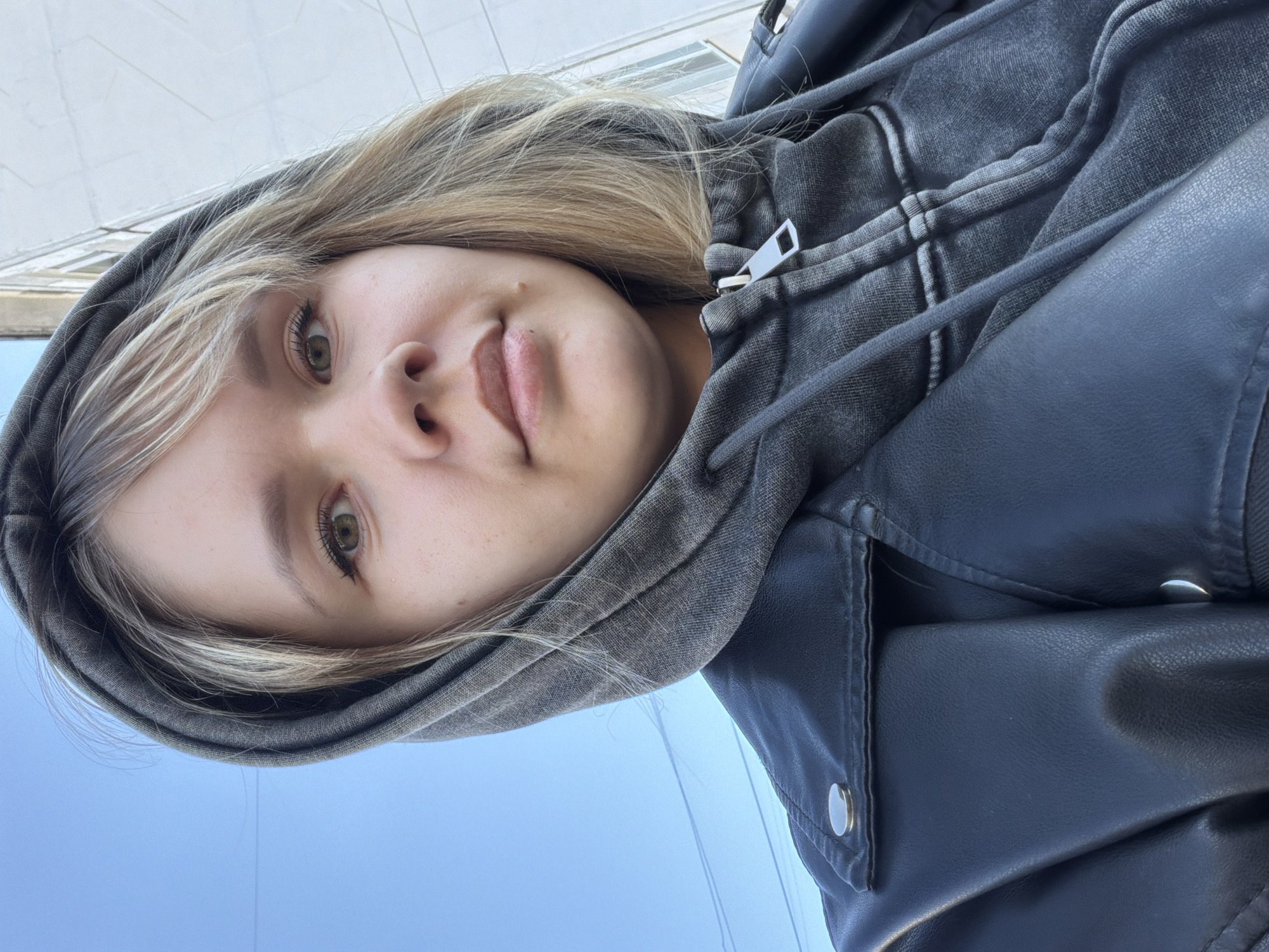A close-up, rotated selfie shows a person with light blonde hair wearing a gray hoodie and a dark leather or leather-like jacket. The background reveals a light blue sky above a light-colored building exterior. The location of the photograph is Unknown, Unknown.