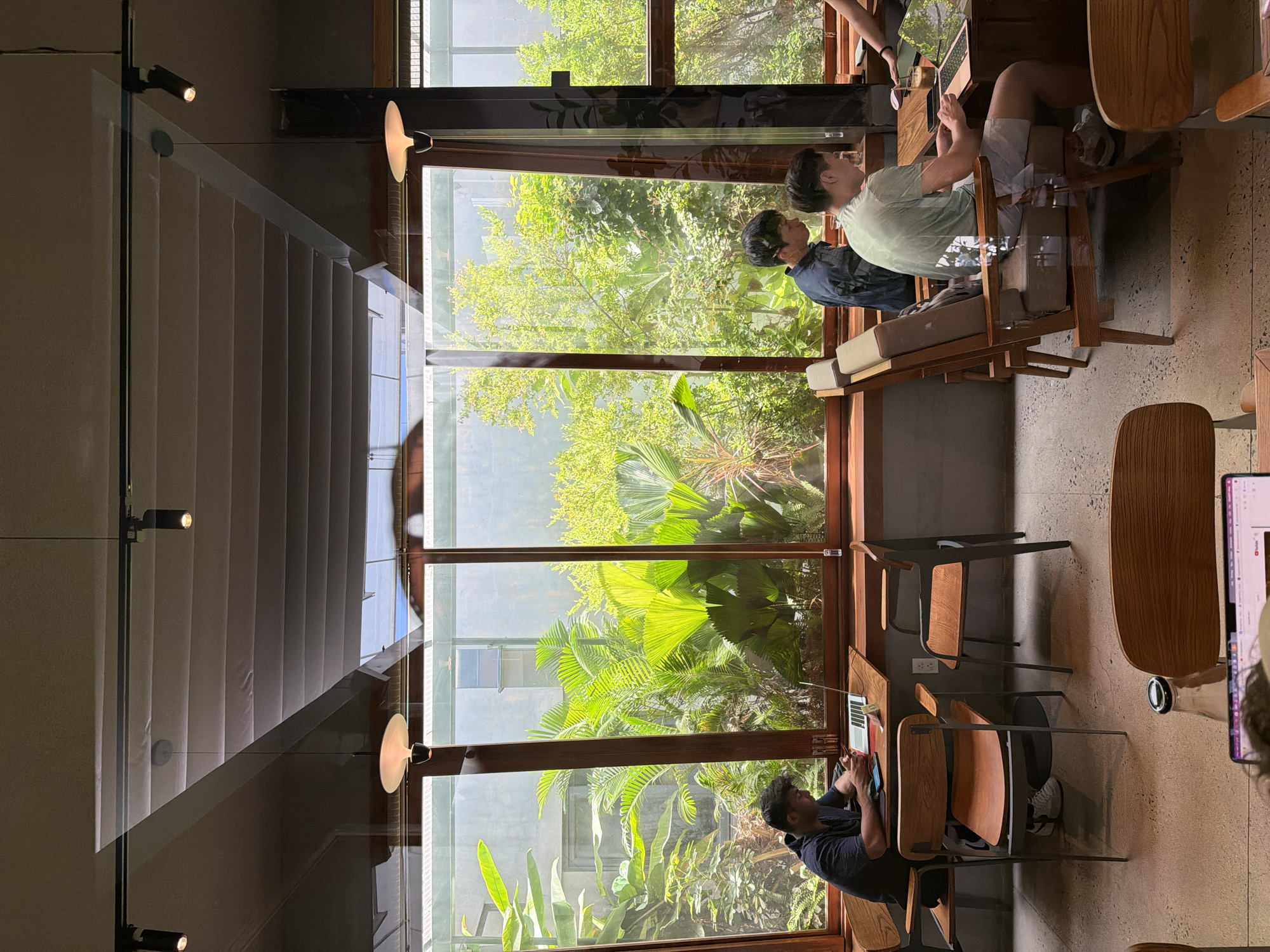 A coffee shop setting in Đà Nẵng, Vietnam, features large windows framing lush green foliage outside. Three individuals are present: one person sits at a table working on a laptop, another person is using a smartphone, and a third person is seated at a table in the background. The interior lighting is a mix of natural light from the windows and artificial light from mounted fixtures. Tables, chairs, and laptops are visible.