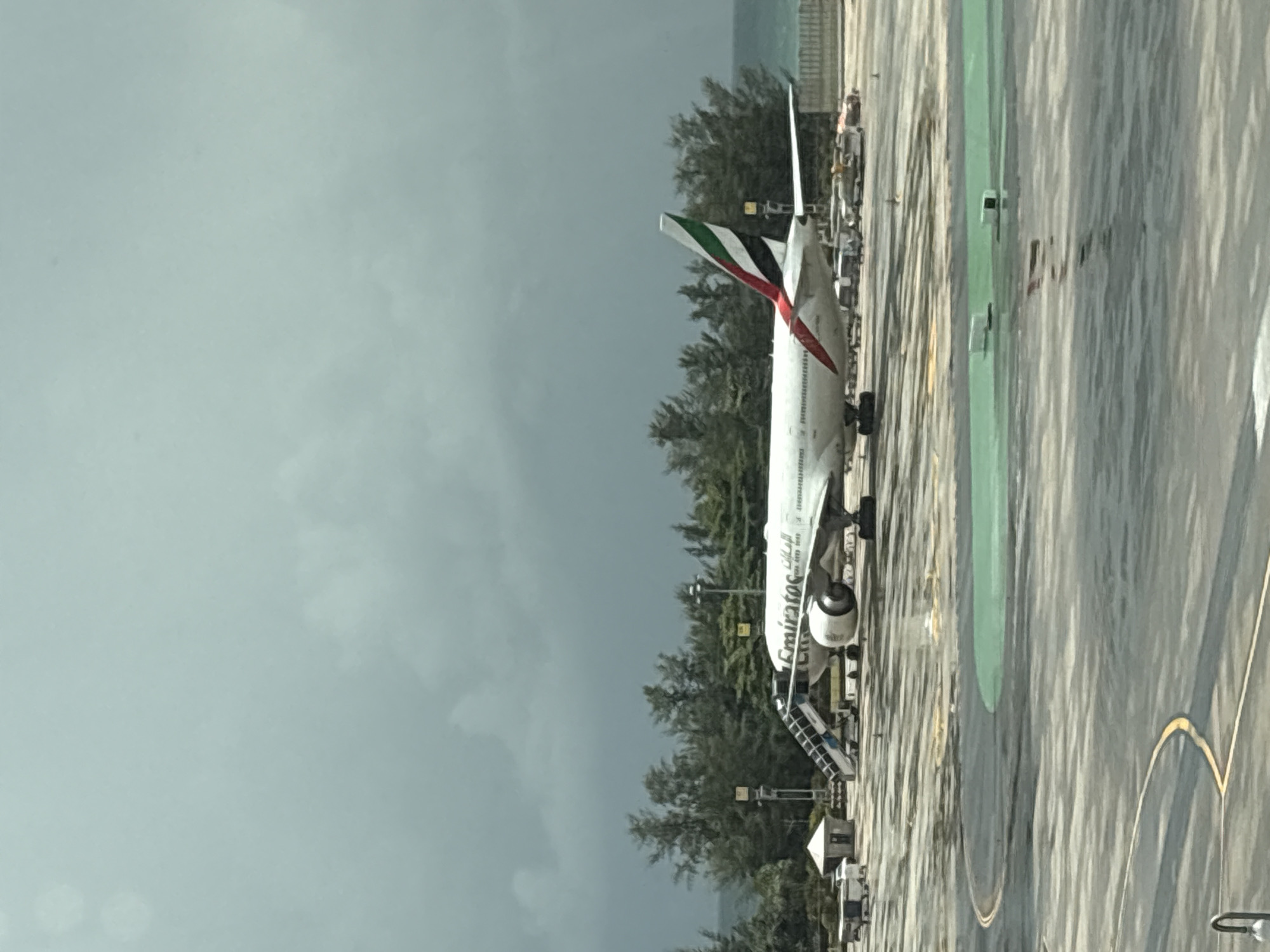An Emirates airplane is parked on a tarmac in Mai Khao, Thailand, under a cloudy sky. The plane is white with red and green accents on its tail fin, and the Emirates logo is visible on its side. A set of stairs is positioned near the front of the plane, connecting it to the ground. Several vehicles, including baggage carts and a small white structure, are also present on the tarmac. In the background, there is a line of trees and the ocean. The tarmac appears wet, suggesting recent rain. The scene is captured from a slightly elevated perspective, possibly from within an airport terminal.