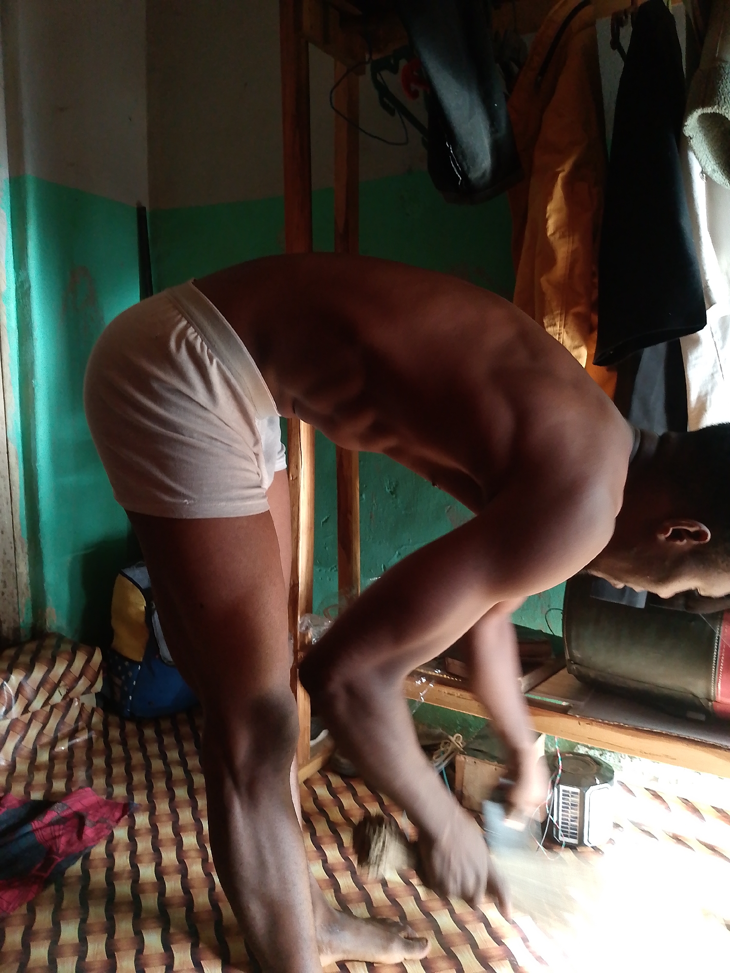 A person is bending over in a cluttered room. The person is dark-skinned and is wearing light-colored underwear. The room has a bed with a brown and beige striped mattress. There is a wooden rack with items hanging on it. The wall is painted green and white. There are various objects on a shelf near the person's hands, including a small black and white appliance with wires. The location is Unknown, Unknown.