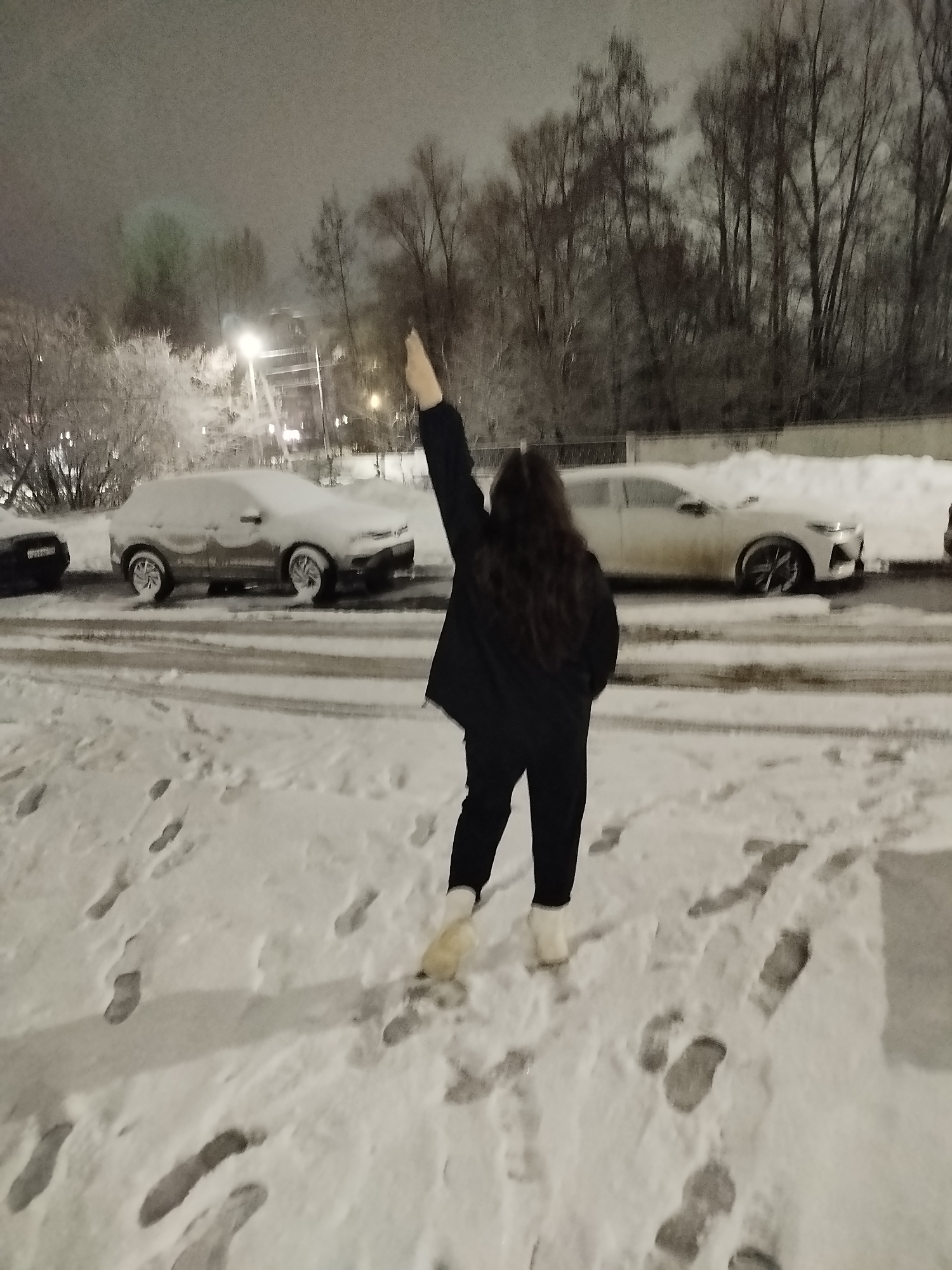 A person stands in the snow in the city of Chaikovsky, Russia, facing away from the camera with their right arm raised. The person has long, dark hair, and is wearing a dark coat, dark pants, and light-colored boots. A car covered in snow is parked nearby, as well as another car that appears white. Bare trees are visible in the background. The ground is covered in snow, with many footprints visible. A streetlight is illuminated in the background. The scene appears to be at night or dusk.