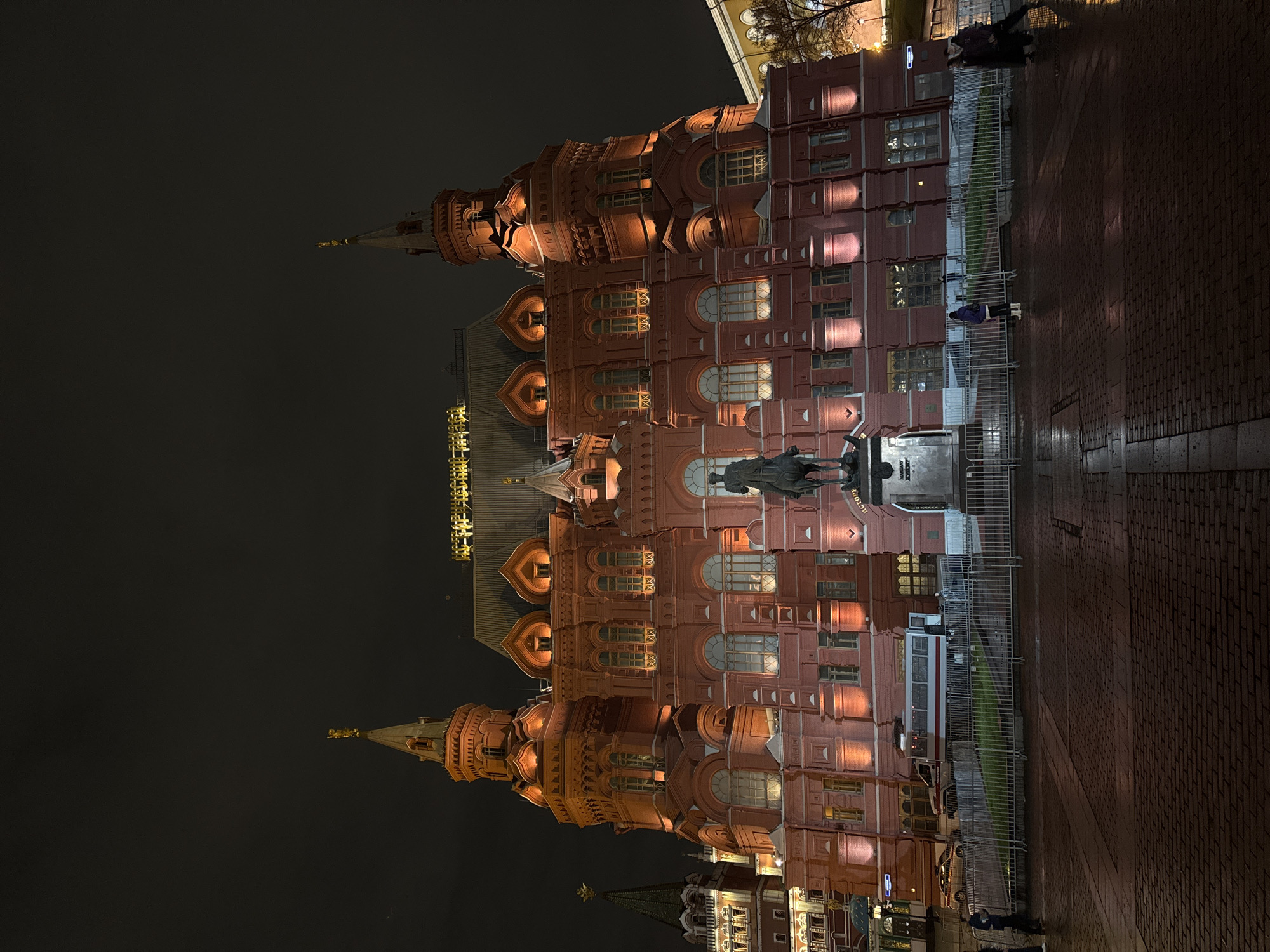 At night, an exterior view captures the State Historical Museum in Moscow, Russia. The red brick facade, illuminated with warm light, contrasts with the dark sky. The building's architecture features decorative details and multiple stories. A statue stands in front of the museum, partially obscured by shadows. A metal fence runs along the building's perimeter. On the right, the pavement is wet. People are visible near the fence. A glimpse of another building can be seen at the bottom.