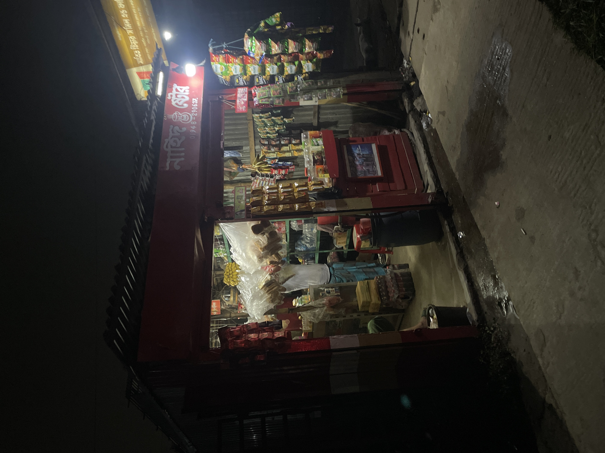 A street view at night in Nasirabad, Bangladesh, shows a brightly lit shop. The shop, named "Nahi Enterprise" in Bengali script, displays a variety of snacks and goods. A man wearing a white shirt and blue pants is inside the shop, likely attending to inventory or customers. Various snacks are hanging from the roof, including chips and biscuits. A light pole is visible and part of the cement street is visible.