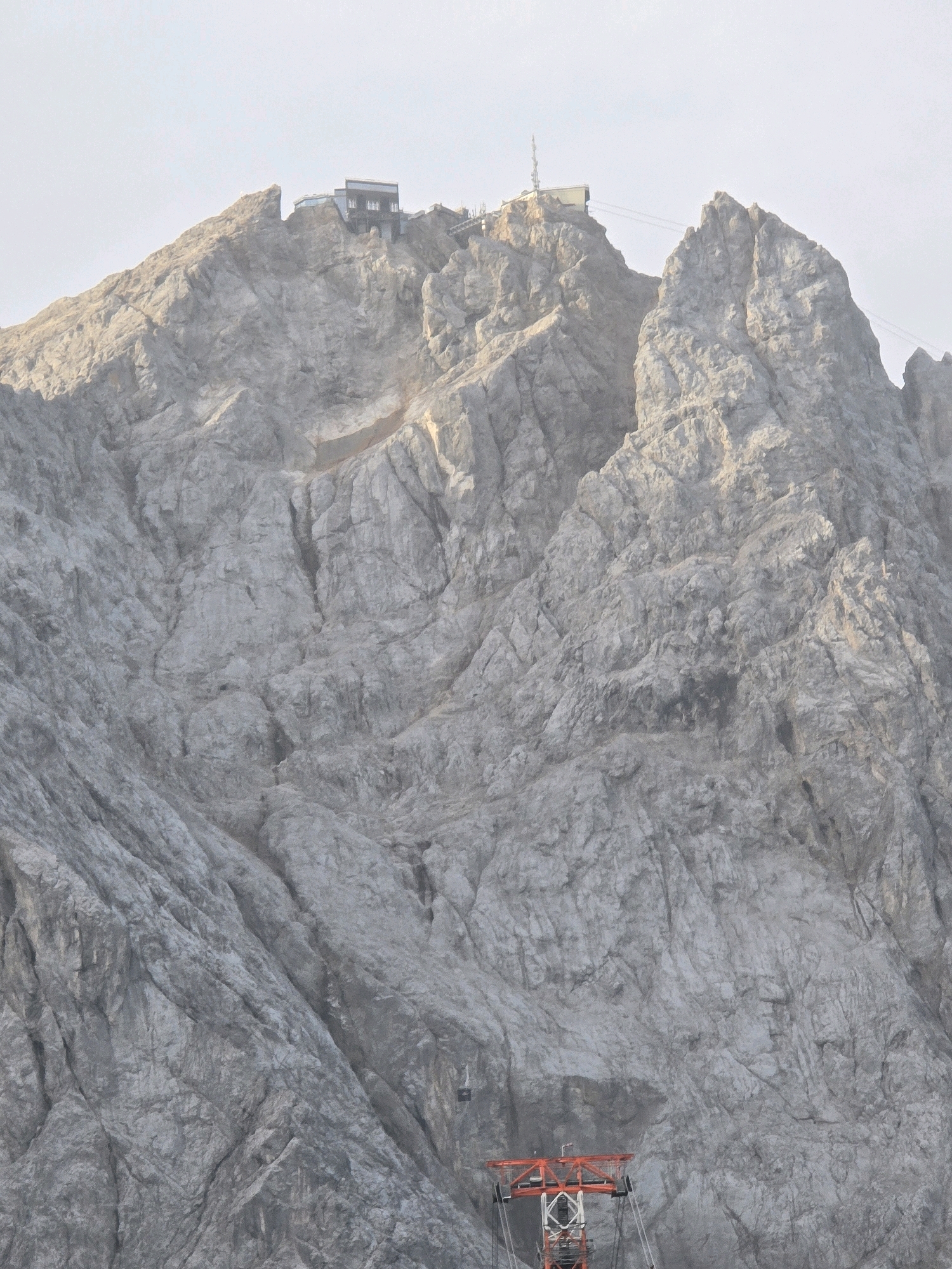 This is a photograph of the Zugspitze mountain in Grainau, Germany, featuring a ski lift and a building on the summit. The image is captured from a distance, looking up at the rocky, mountainous terrain. The sky is overcast and pale. In the foreground, the top of an orange ski lift structure is visible. Further up the mountain, a building with a dark roof and windows is perched on a rocky outcrop. There are no people visible in the image. The environment is rugged and mountainous, with visible rock formations and sheer drops.