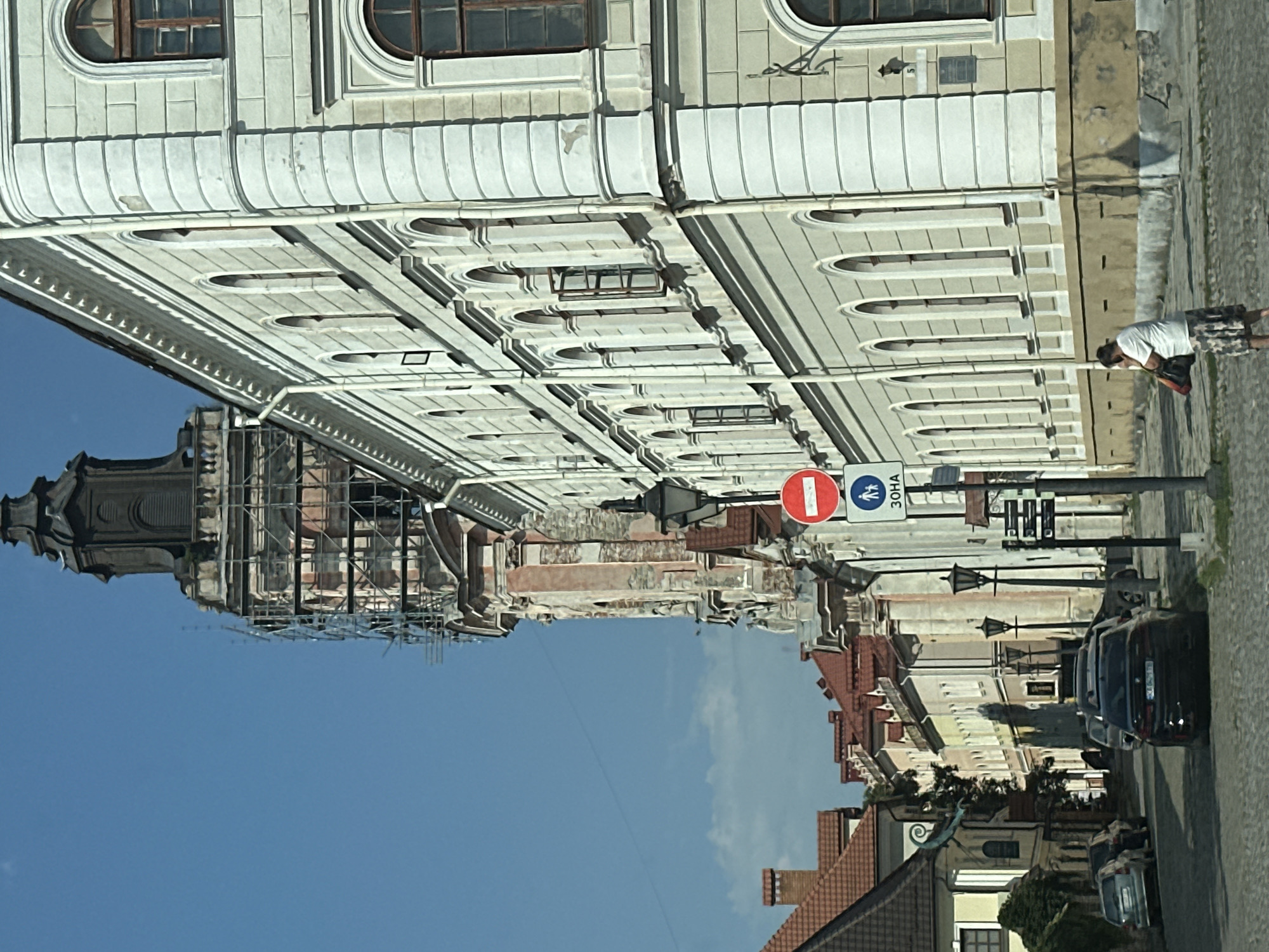 The image shows a street scene in Kamianets-Podilskyi, Ukraine, with a prominent view of the Old Town Hall tower, which is partially covered in scaffolding. To the right of the tower is a light-colored, multi-story building with numerous arched windows and decorative architectural elements. A street lamp stands on the right side of the road, accompanied by traffic signs, one circular red and white sign indicating prohibition, and a rectangular sign with pedestrian icons and the Ukrainian text "ЗОНА" (ZONA). Several cars are parked on the cobblestone street. In the foreground, a woman wearing a white t-shirt and a patterned skirt is walking, carrying a bag. The sky is clear and blue with some scattered clouds. The street is paved with cobblestones, and there are other buildings visible further down the street, some with red tile roofs.
