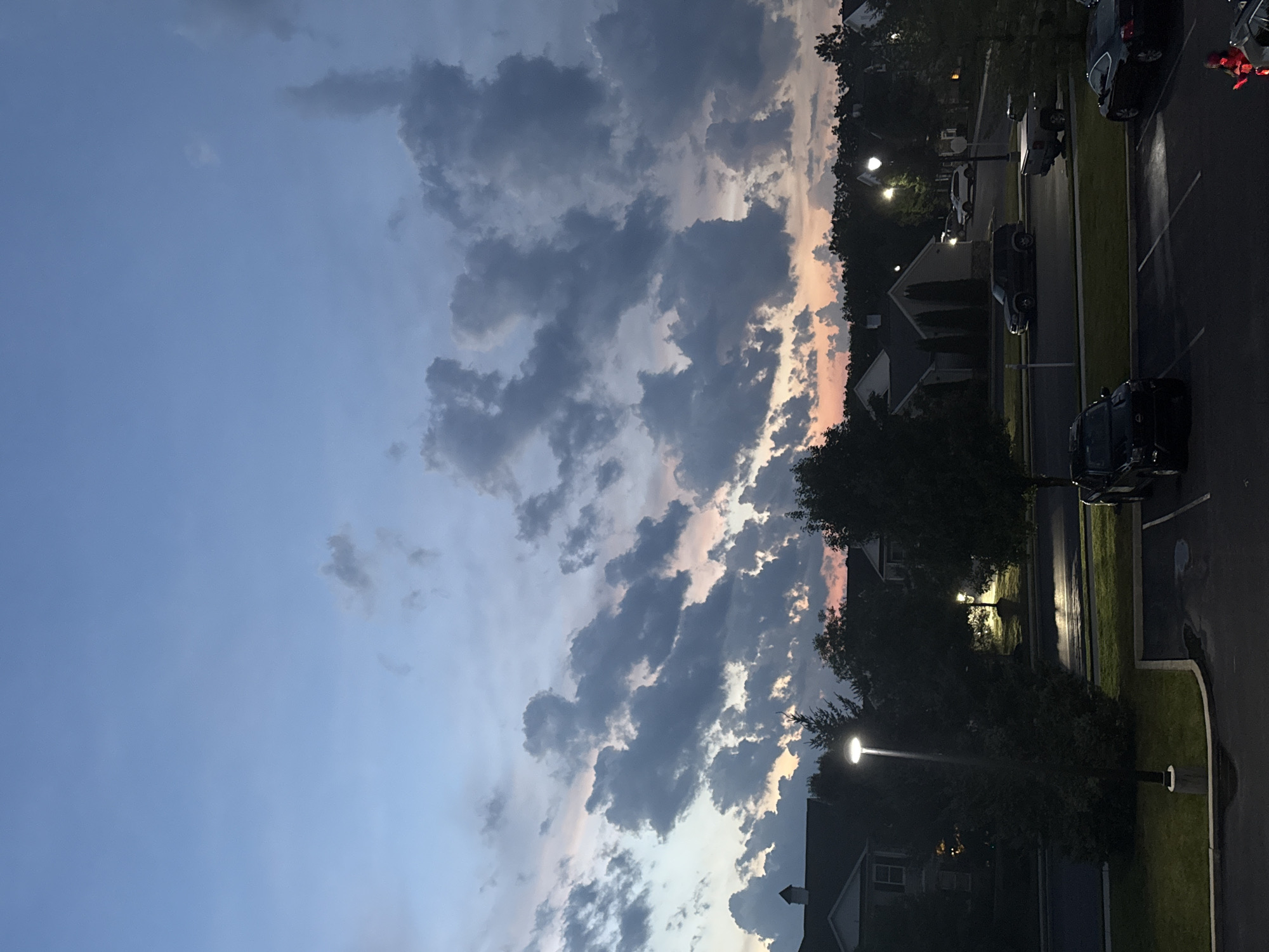 An overhead view shows a twilight scene in Allen Township, United States of America. The sky transitions from a pale blue to a soft pink and orange near the horizon, where it is dotted with dark, cumulus clouds. Below the sky, a suburban landscape is visible. Several houses with dark roofs and light-colored siding are nestled amongst dark green trees. A street runs through the middle of the scene, with a few parked cars, including a black SUV and a silver pickup truck. A streetlamp illuminates a patch of wet pavement and grass. The foreground consists of a dark parking lot with a few more vehicles, including a black SUV and a black sedan. A red object, possibly a person in a costume, is visible in the lower right corner of the image.
