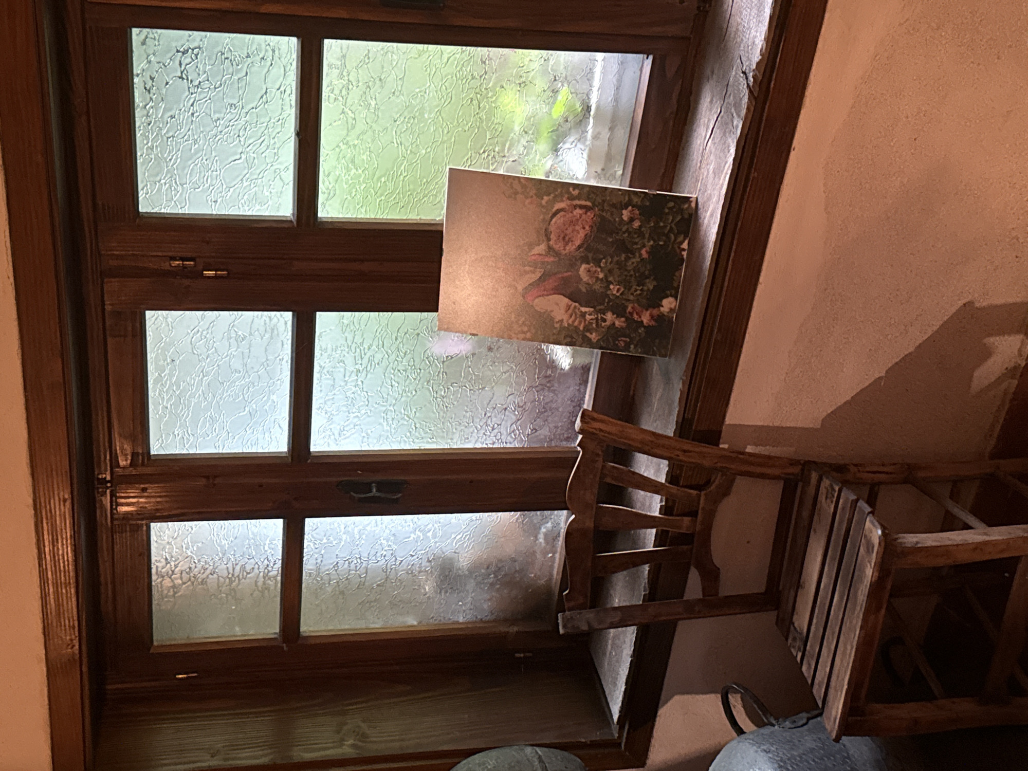 This image captures an indoor scene in Tarnicheni, Bulgaria, centered on a wooden window. The window features a dark brown frame and multiple panes, most of which contain textured, patterned glass that obscures the exterior view. The panes on the right reveal blurred green foliage outside. Visible window hardware includes hinges and a latch. A weathered wooden window sill, showing cracks, extends beneath the window. A wooden chair with a slatted back and seat is positioned to the left of the window. On the right side of the window sill, a rectangular print or artwork leans against the frame, depicting a figure, likely a child, wearing a red vest and holding a basket, surrounded by pink roses. The wall below the window and to the left of the chair is textured and light orange. A metallic or grey object is partially visible on the far left. Natural light from the window illuminates the scene, casting shadows within the room.