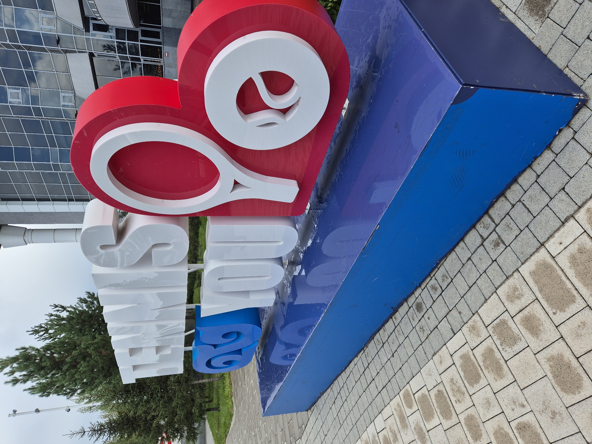 The image shows a large, multi-colored outdoor sculpture in front of a modern building. The sculpture is composed of the words "TENNIS" in large white 3D letters, followed by "ES" in blue, and then "YOU" in white. The letters "TENNIS" are rendered in a sleek, modern font with a slightly reflective surface. The blue letters "ES" have a swirling, wave-like design. The white letters "YOU" are also in a prominent 3D style, and the "O" in "YOU" is integrated with a red and white heart shape that resembles a tennis racket. The entire sculpture is mounted on a rectangular blue base.
The background shows a large, glass-fronted building, likely an arena or stadium, with a modern architectural style. To the left of the sculpture, there are several tall green evergreen trees and a paved walkway. The ground in front of the sculpture is paved with light-colored interlocking stones, which appear damp, suggesting recent rain or a wet surface. The overall impression is that of an outdoor public art installation, likely at a sports venue. The location is identified as Astana, Kazakhstan.