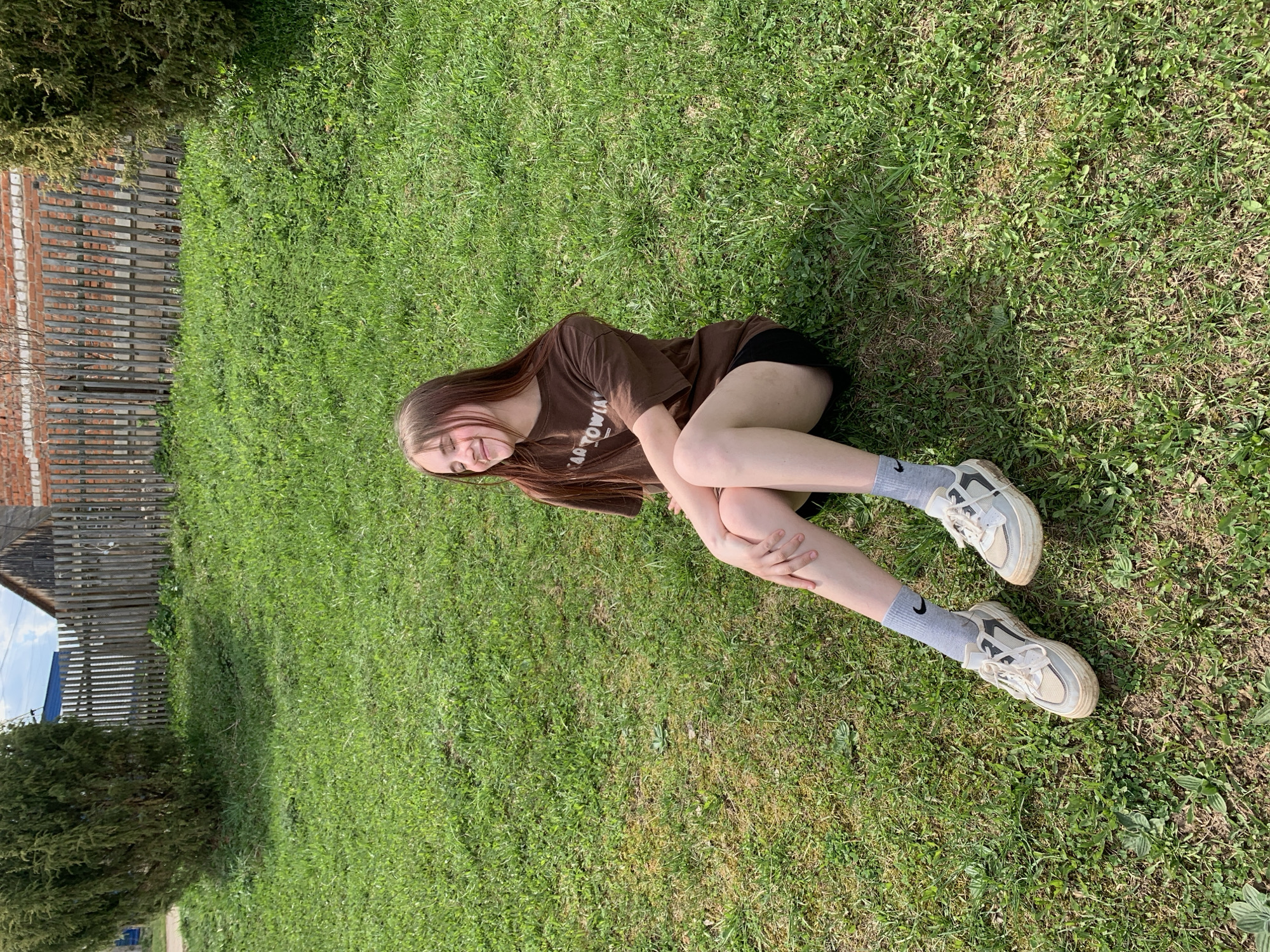 A young woman is sitting on a grass lawn. She is wearing a brown t-shirt, black shorts, grey socks and dirty white shoes. She is facing the camera and smiling. There is a brick and wood fence in the background with a green tree and grass. The location is assumed to be Фоки, Russia.