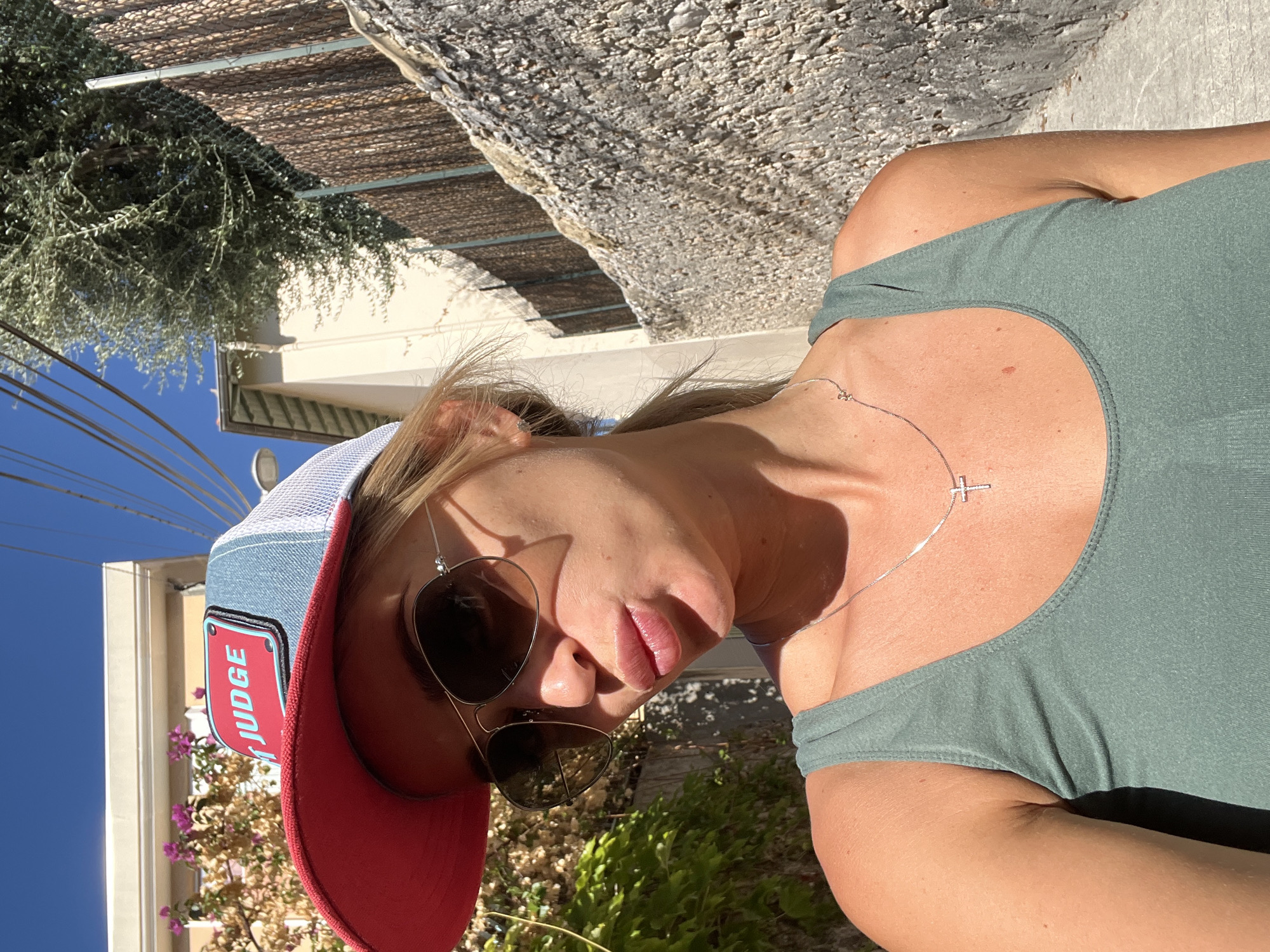 A woman, wearing a red and blue trucker hat with a "JUDGE" patch, aviator sunglasses, and a green tank top, takes a selfie outdoors in Nice, France. She is posing with her lips puckered. The background features a stone wall, vegetation, and buildings under a bright blue sky.