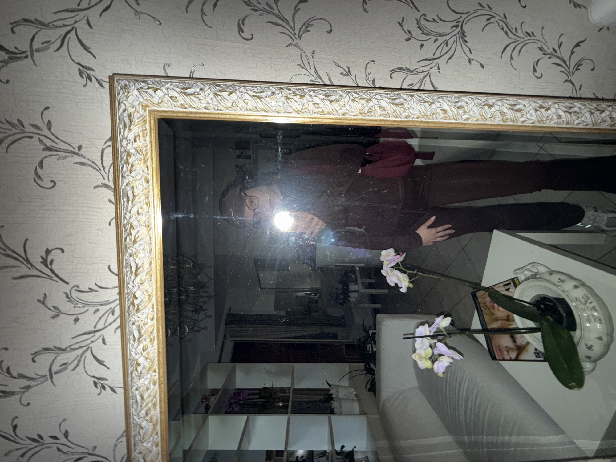 A slightly angled, medium shot captures a reflection in a large, ornate mirror hanging on a wall covered in a light-colored wallpaper with dark, swirling floral patterns. The mirror has a white and gold-colored frame with a detailed, leafy design. The reflection shows a person wearing glasses and a dark-colored outfit with a red bag on their shoulder, holding a phone with the flash on, presumably taking the photo. The reflection also reveals a glimpse of the room, including a white shelf with various items, a white sofa, and an orchid plant in a decorative pot on a white table. The scene is dimly lit.
There is no information available to place this scene in the city of Sanremo, Italy.