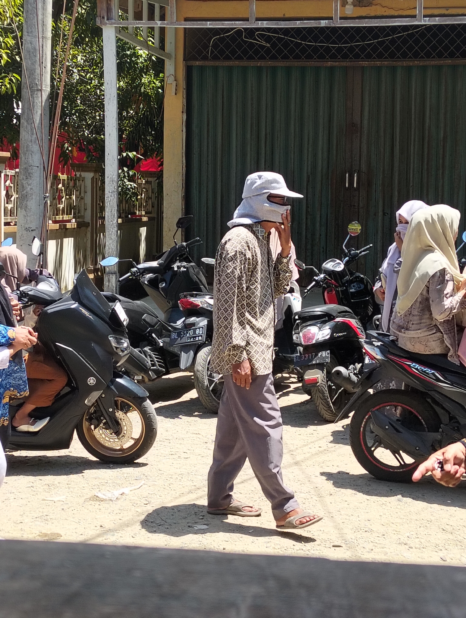 In Teungoh, Indonesia, a man wearing a light-colored wide-brimmed hat and a patterned shirt walks across a dusty outdoor area, with his face partially covered by a mask or cloth. He is right of center. Behind him, several motorcycles are parked. To the far right, a person's hand is visible, pointing towards something off-camera. Two women wearing hijabs are standing near the motorcycles, to the right of the walking man. The background features a building with a large green corrugated metal door. The scene appears to be on a sunny day, casting distinct shadows on the ground.